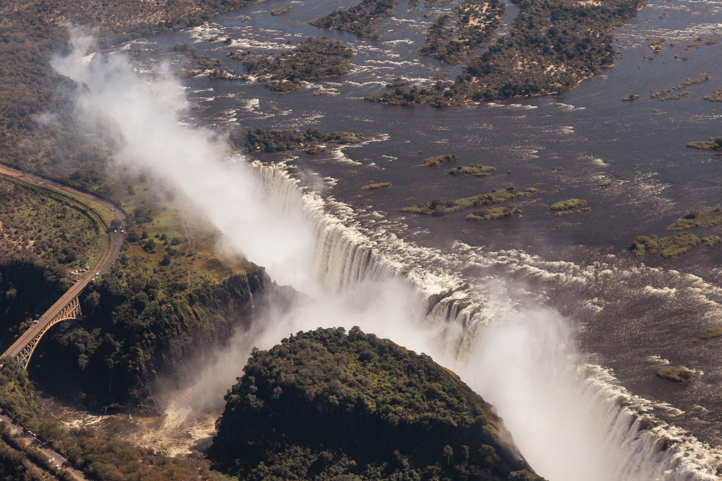 Aerial view of Victoria Falls with the Zambezi River pouring into a canyon, mist rising in a tall curtain, rainforest and road on the cliff edge