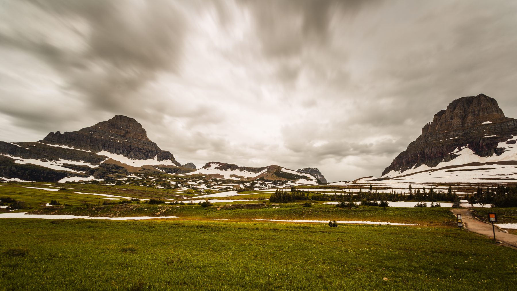 Snow-streaked peaks above a green meadow at Logan Pass under heavy cloud