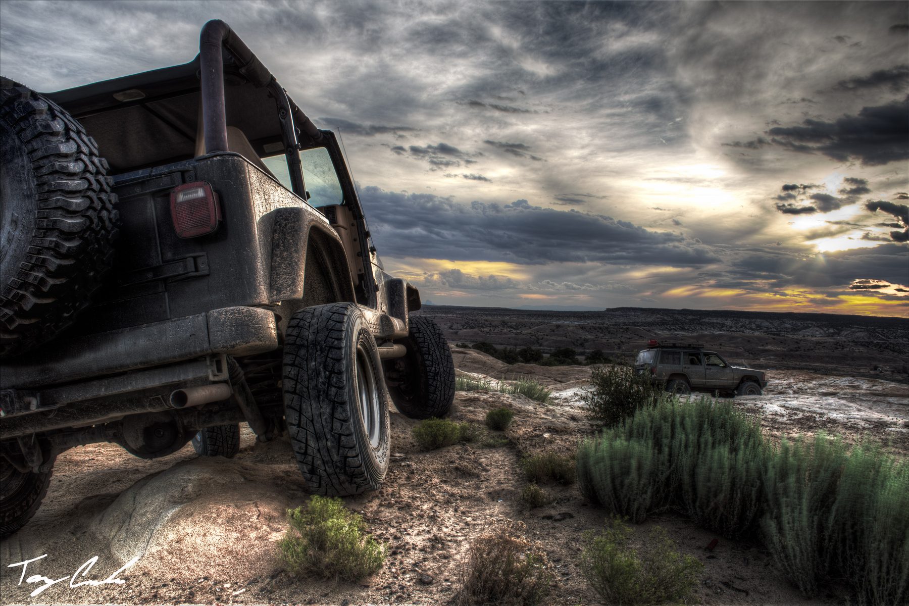 A mud-covered Jeep tipped over a slickrock ledge under a dramatic evening sky