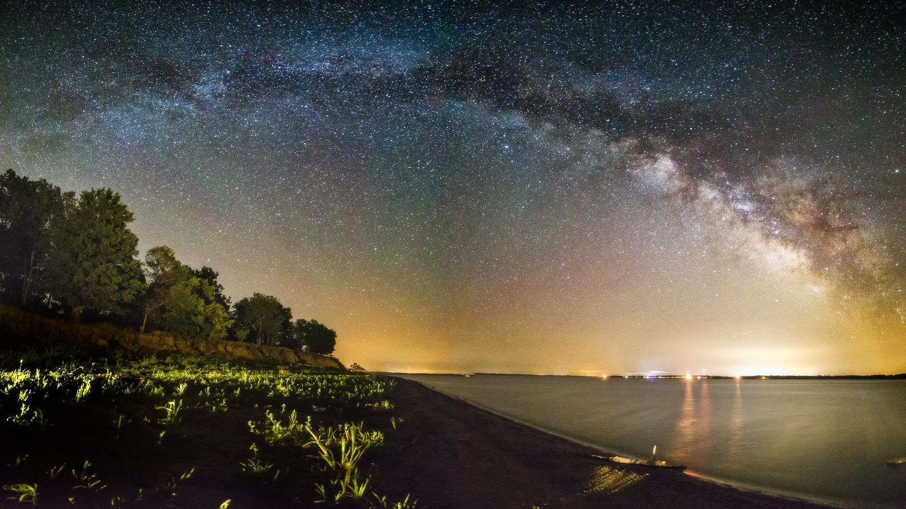 The Milky Way arching over a dark bay with distant town lights on the far shore