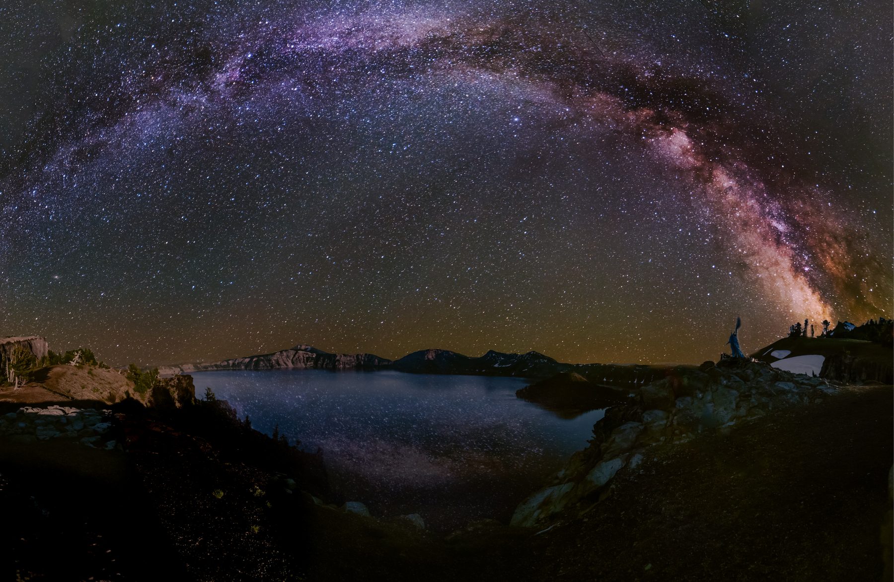 The Milky Way arching over Crater Lake with a figure standing on the rim