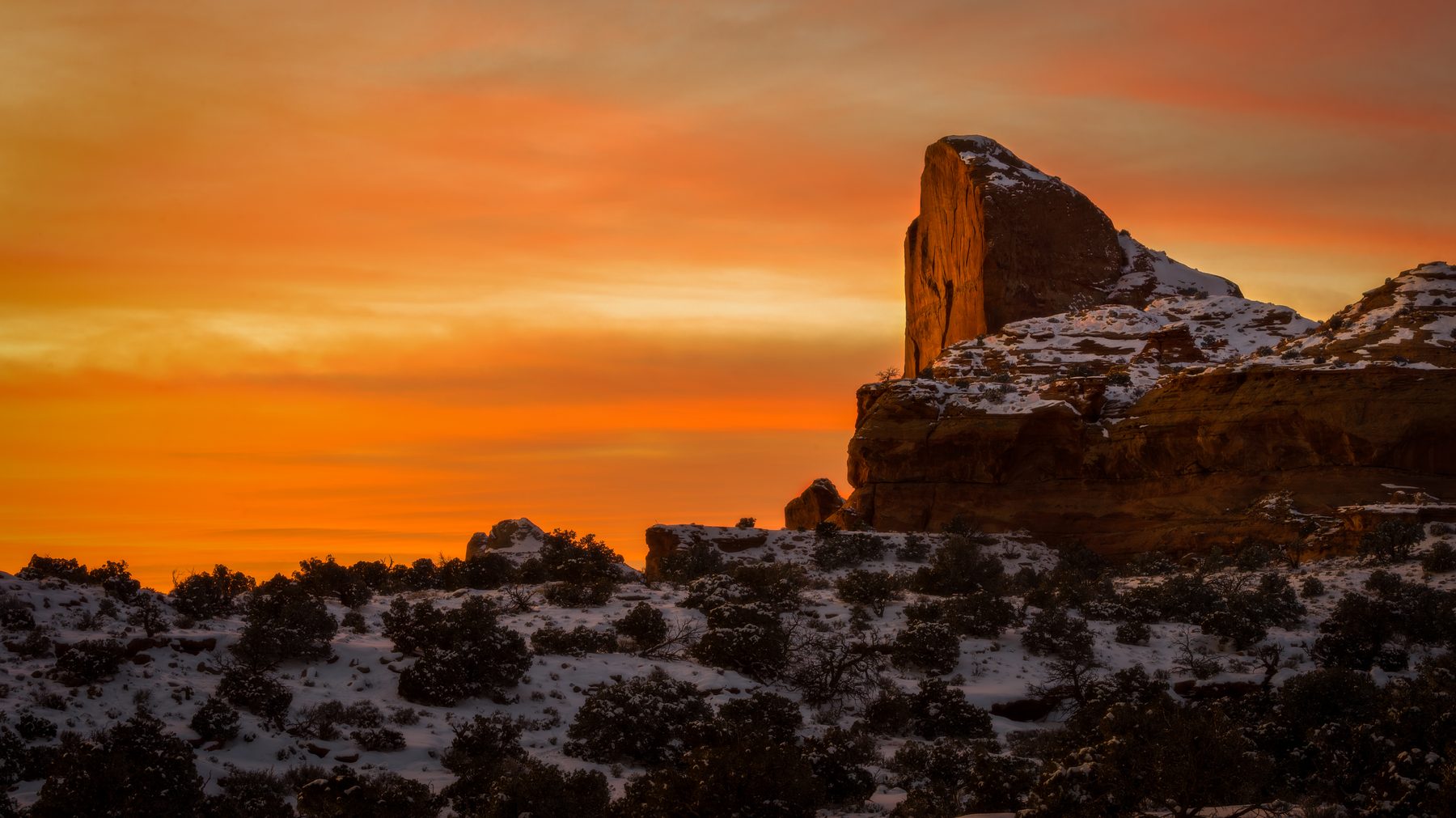 A sandstone spire against a burning orange sky over snow-patched desert