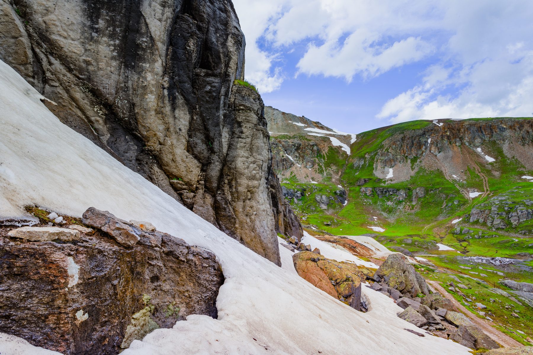 A steep snowfield running out below a dark cliff into a green alpine basin
