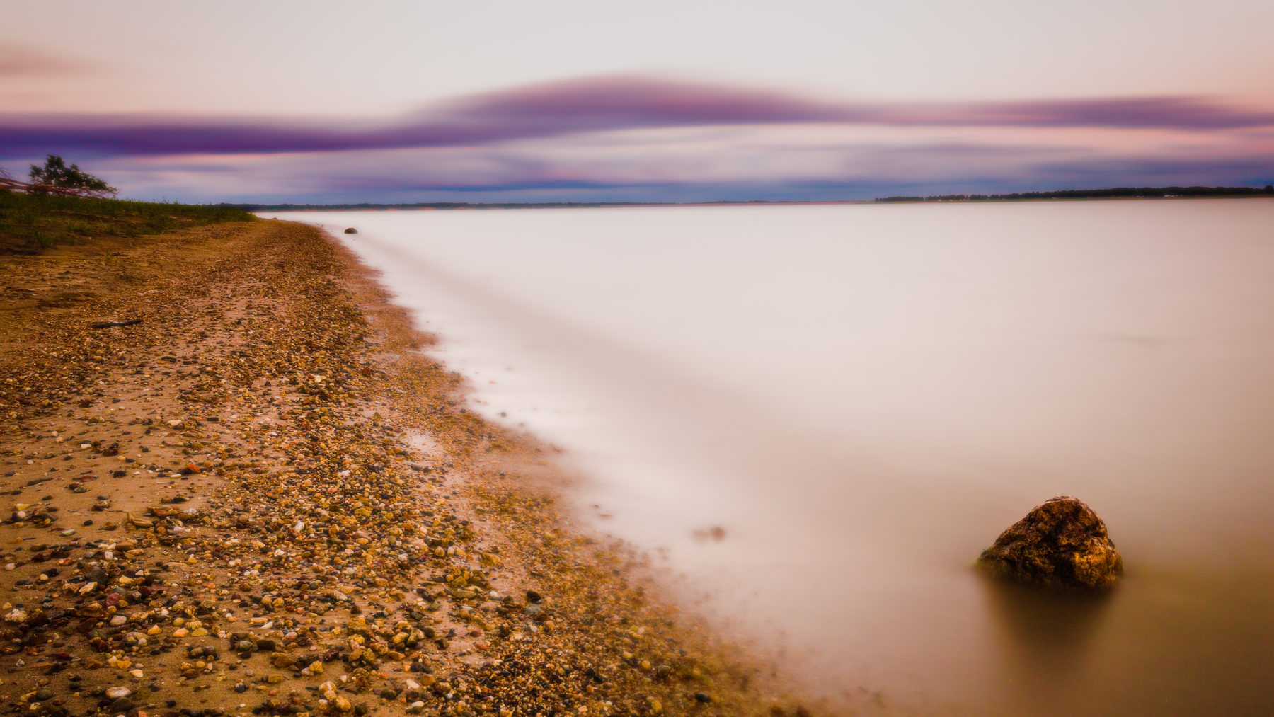 A long exposure of a pebbled shoreline with a single rock in glassy water