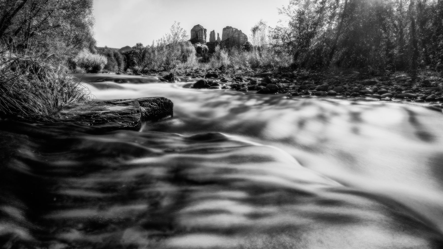 A black and white long exposure of a creek running under red rock buttes