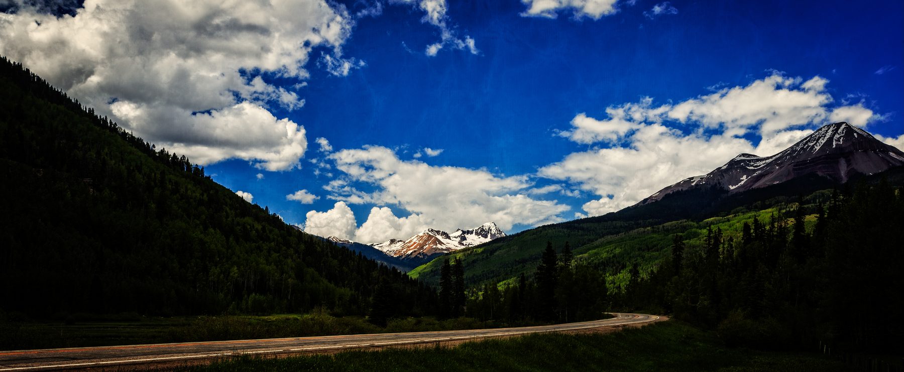 A wide view along a mountain highway toward snow-streaked peaks and cumulus