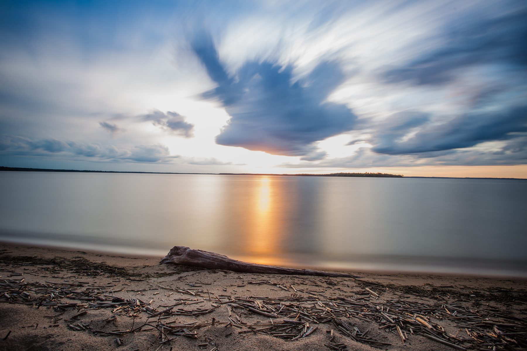 Wispy cirrus cloud streaking over a still lake with driftwood on the sand