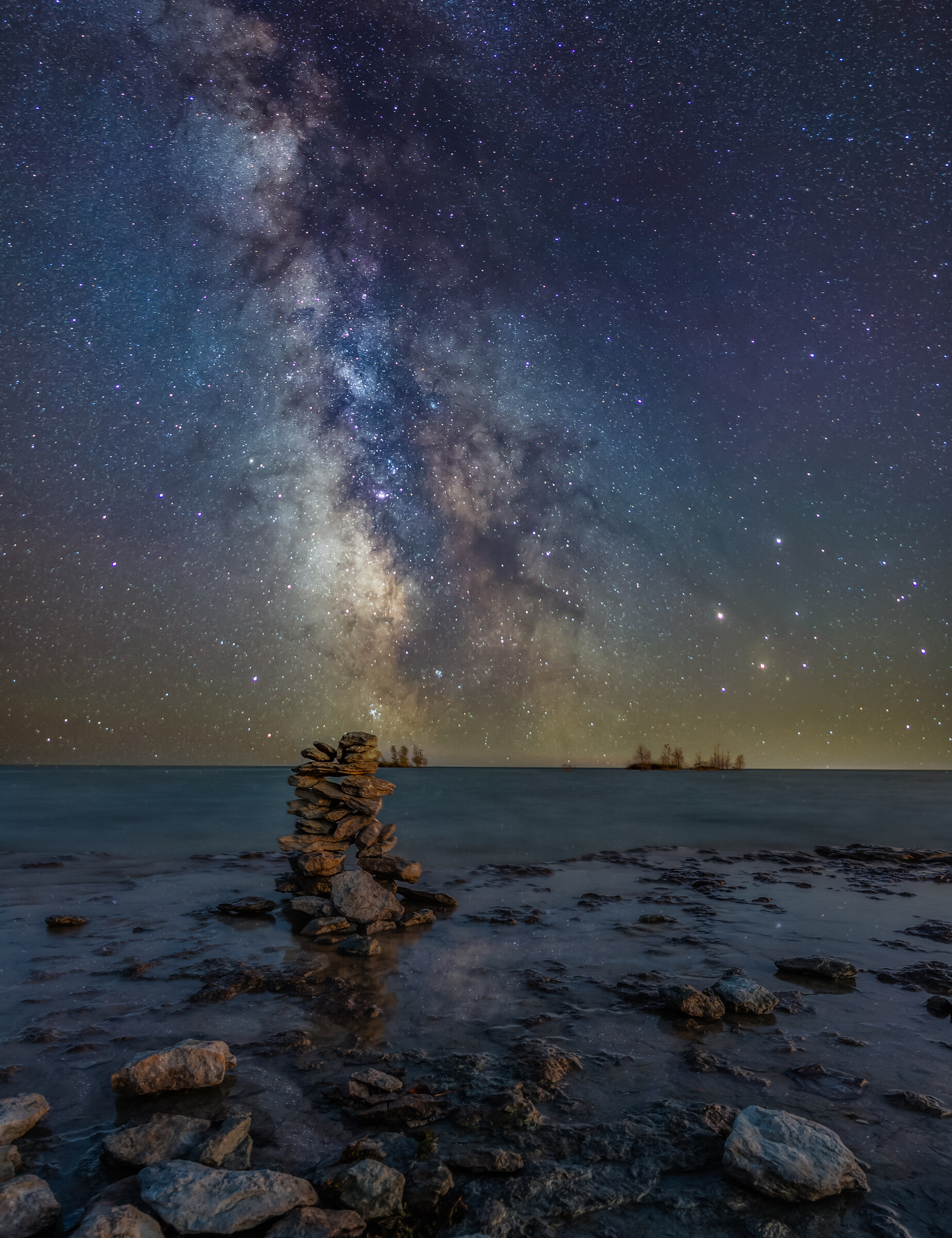 Vertical view of the Milky Way core rising above a stacked-stone cairn on a rocky lake shore, smooth water and a distant treeline