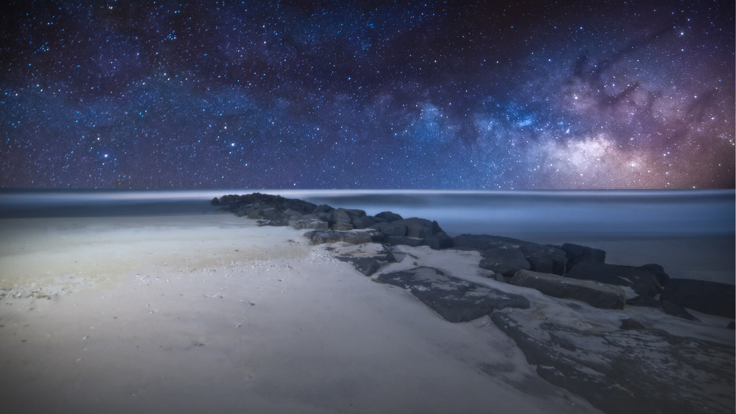Wide landscape of an Atlantic beach at night, a rock jetty extending into the ocean and the Milky Way core visible low on the right side of the sky