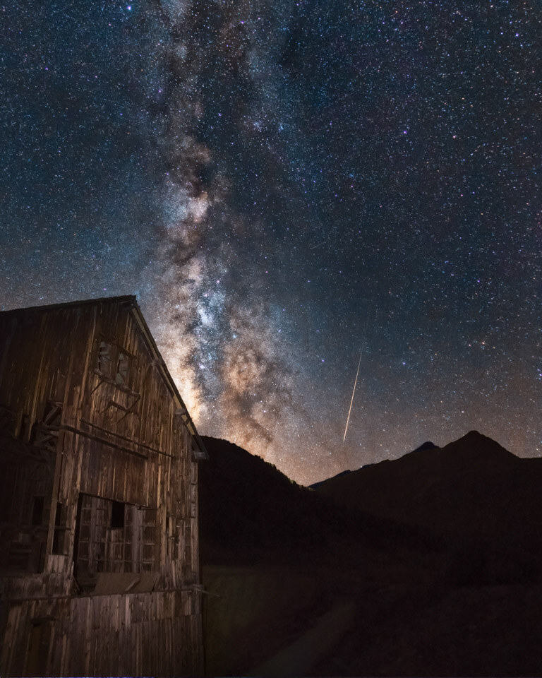 Vertical scene of an old wooden mining structure in a mountain valley at night, the Milky Way core rising above it and a meteor streaking down on the right