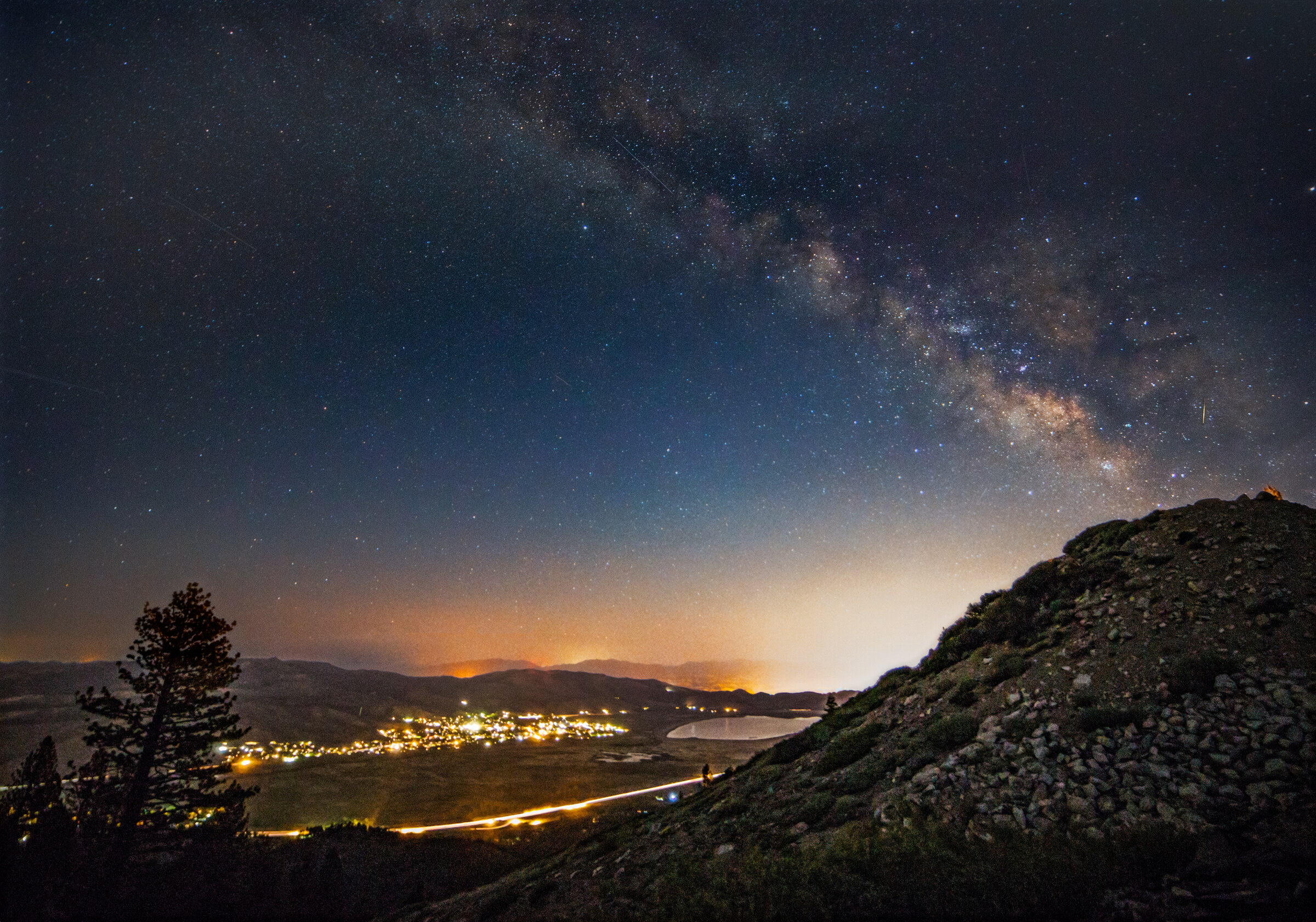 Wide landscape of a mountain valley at night, a small town's lights glowing in the basin and the Milky Way arching overhead in a deep blue sky