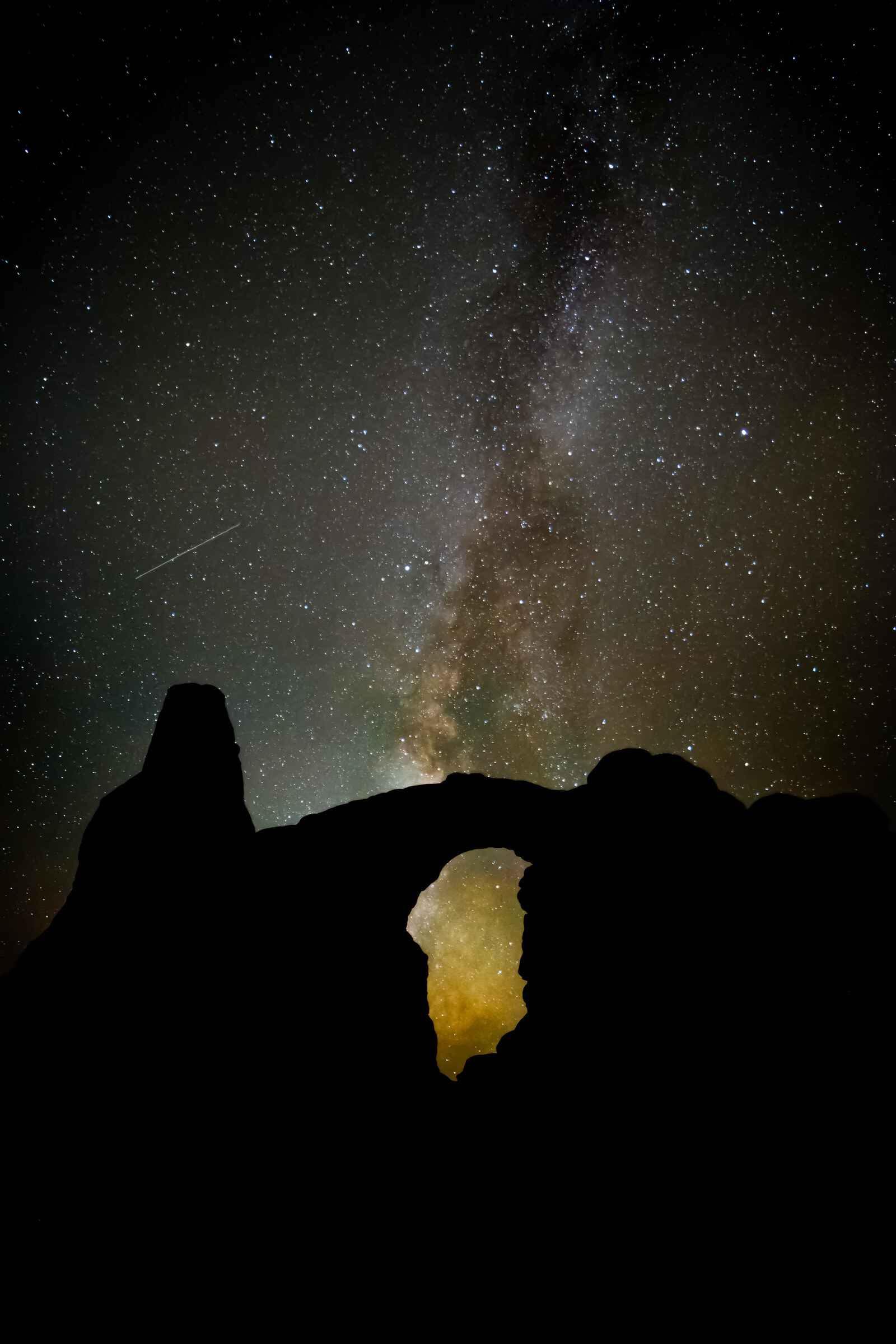 Vertical view of the Milky Way core framed by a silhouetted sandstone arch, faint glow of distant city light visible through the opening
