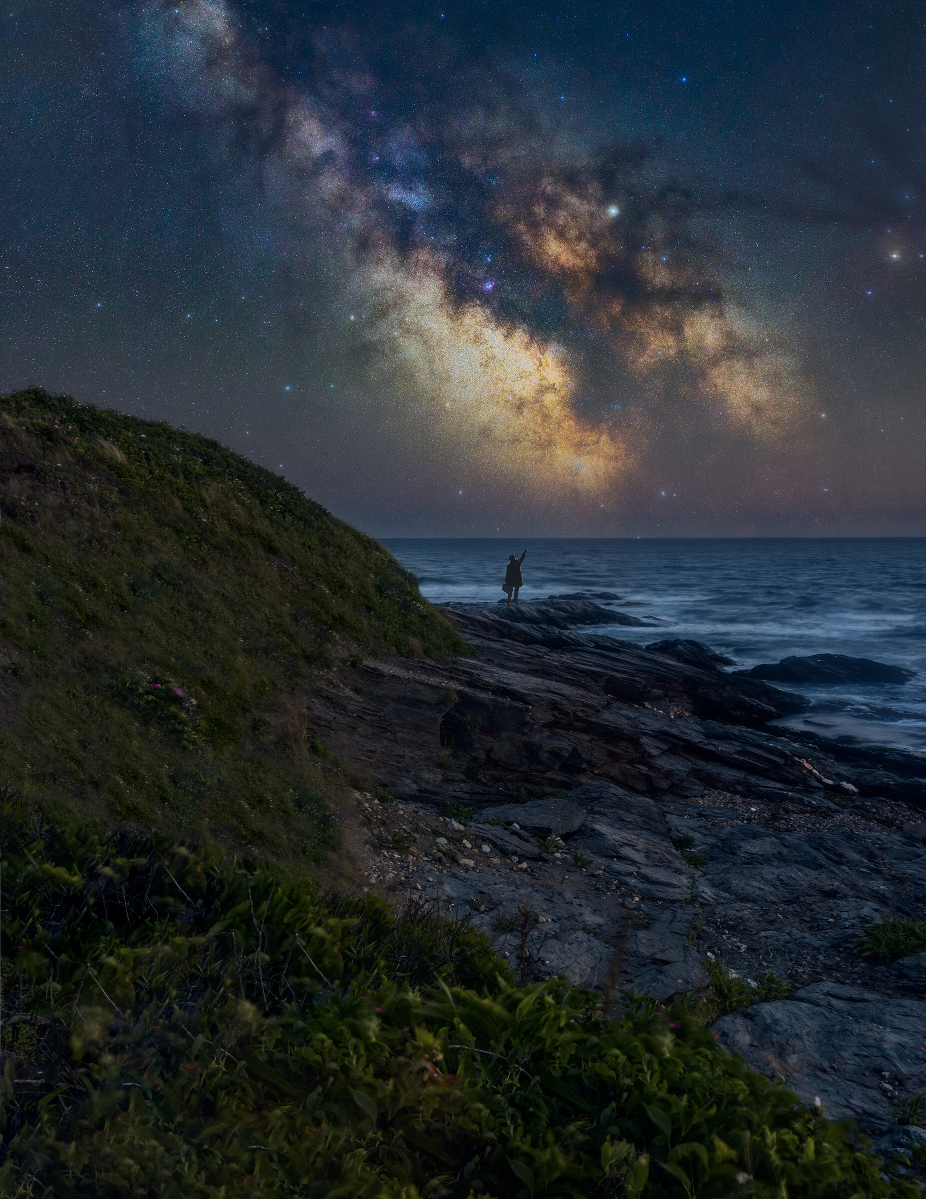 Vertical view of the Milky Way core rising over a rocky New England coastline at night, a small figure of a person standing on the cliff edge for scale
