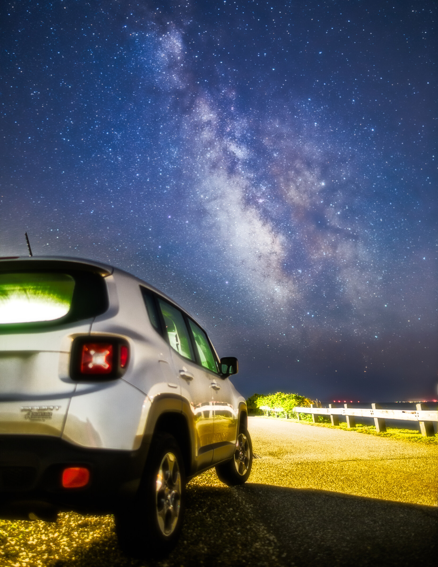 Vertical view of a silver Jeep Renegade parked on a paved coastal pullout at night, the Milky Way core rising behind it in a deep blue sky