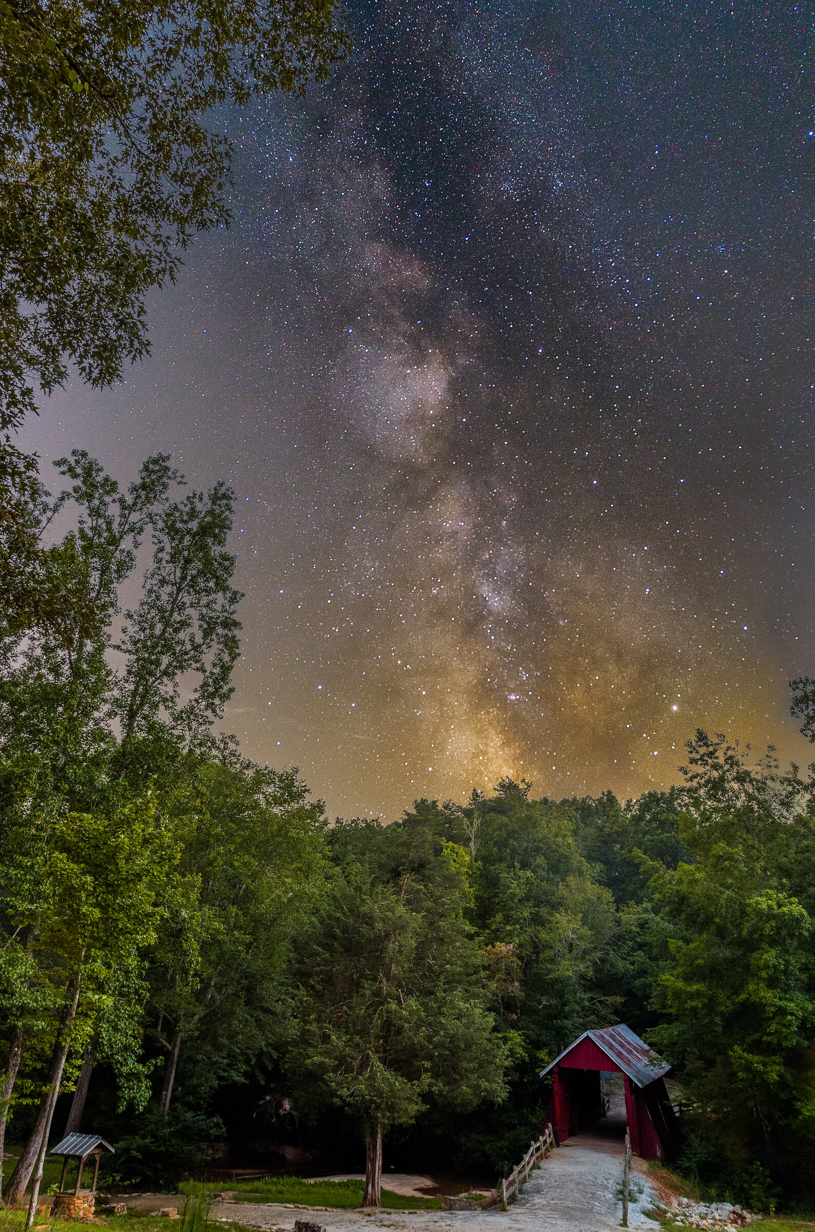 Vertical view of the Milky Way core rising above a red covered bridge nestled in dark trees