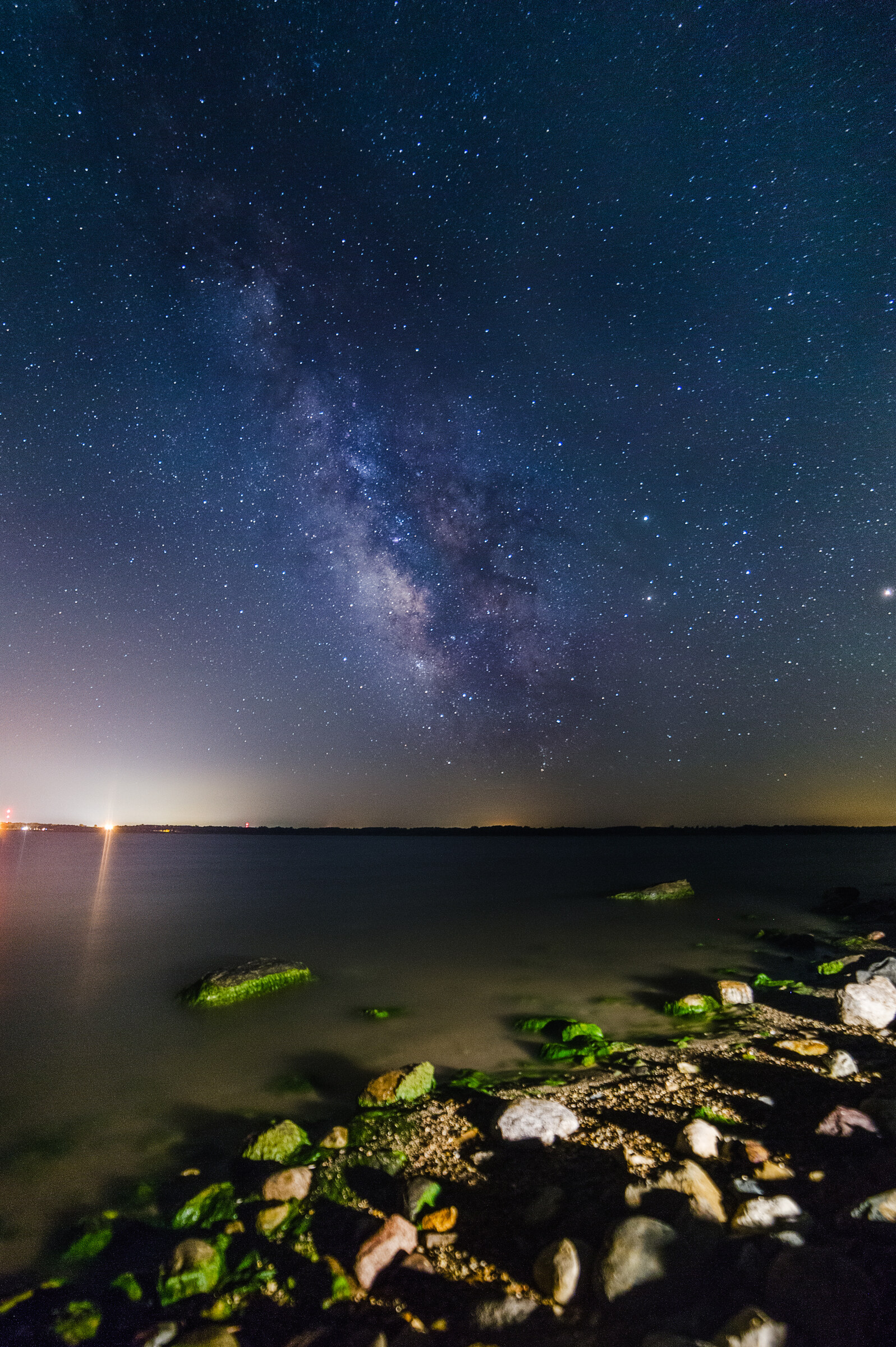 Vertical seascape at night with the Milky Way core in the center of the sky, seaweed-covered rocks in the foreground, a small distant lighthouse glowing yellow on the left horizon