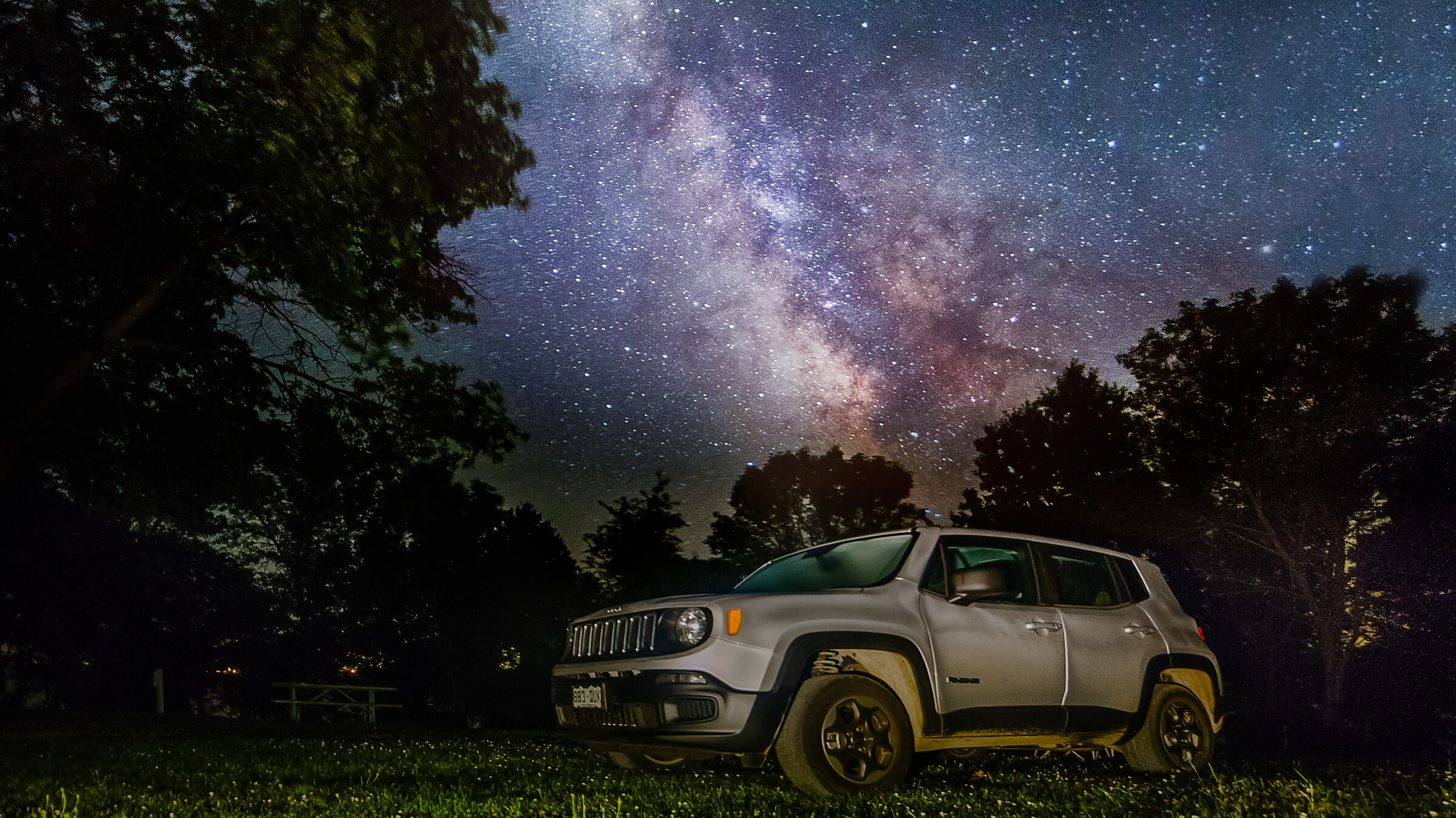 Wide landscape view of a Jeep parked in a grassy field at night, surrounding trees framing the scene, the Milky Way core rising overhead