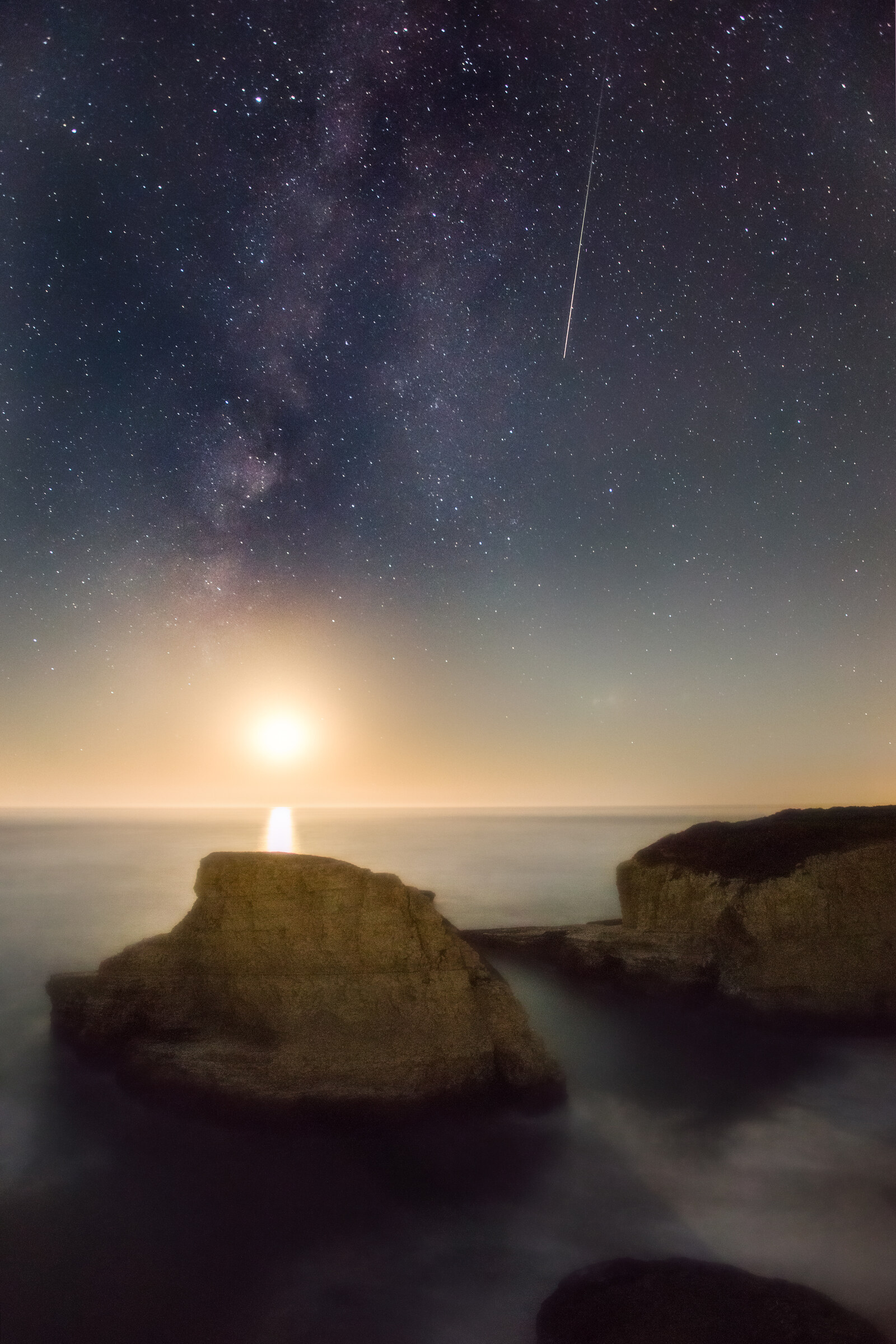 Vertical seascape at night with the Milky Way overhead, the moon setting into the Pacific with a long reflection on still water, a meteor streaking from upper right, and fin-shaped sea stacks in the foreground