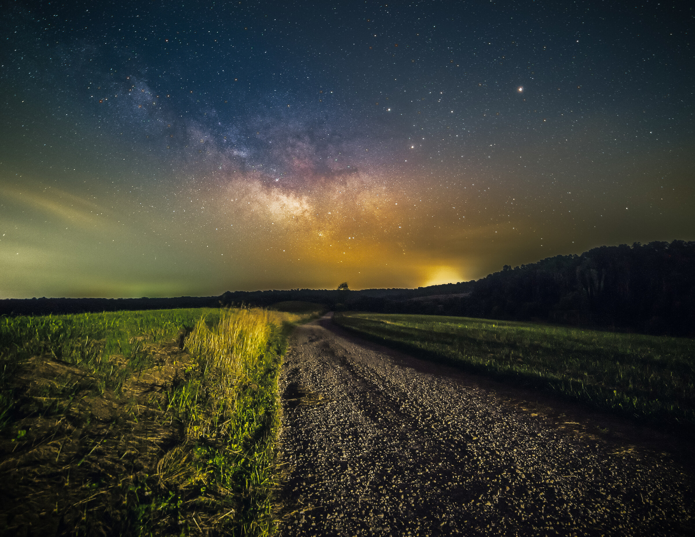 Milky Way core rising above a rural gravel road that cuts through farmland, grass and trees on either side, faint distant light pollution on the horizon