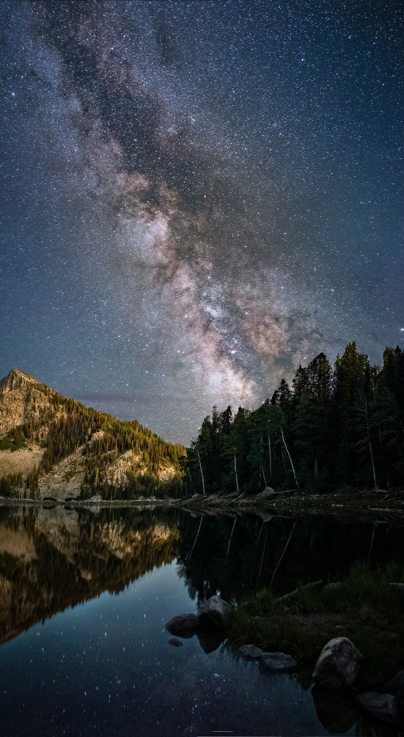 Vertical view of the Milky Way core perfectly reflected in a still alpine lake, mountain to the left and conifer trees on the far shore