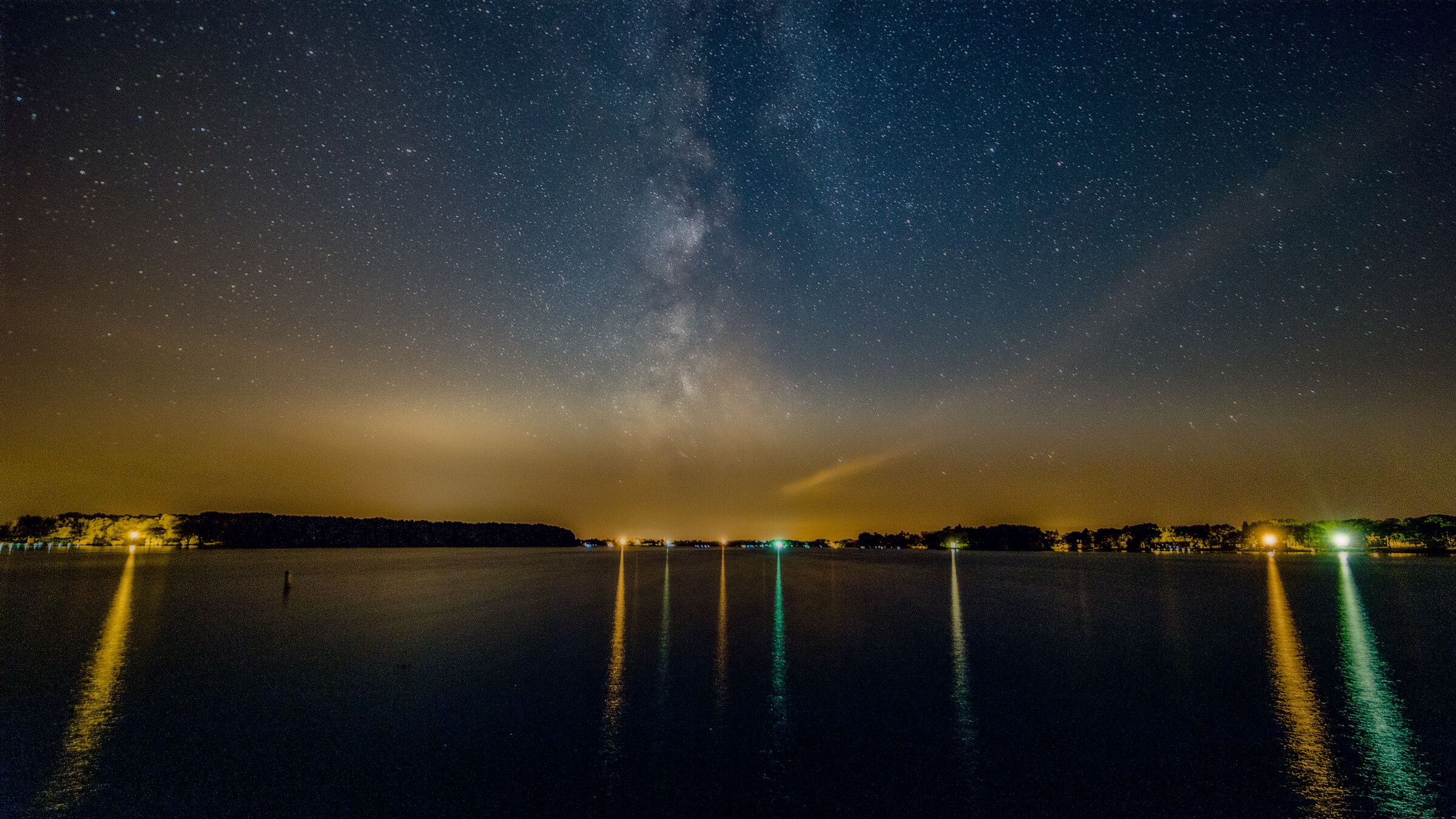 Landscape view of the Milky Way over a calm lake or harbor, multiple yellow and green lights from the far shore reflecting in long stripes on the water