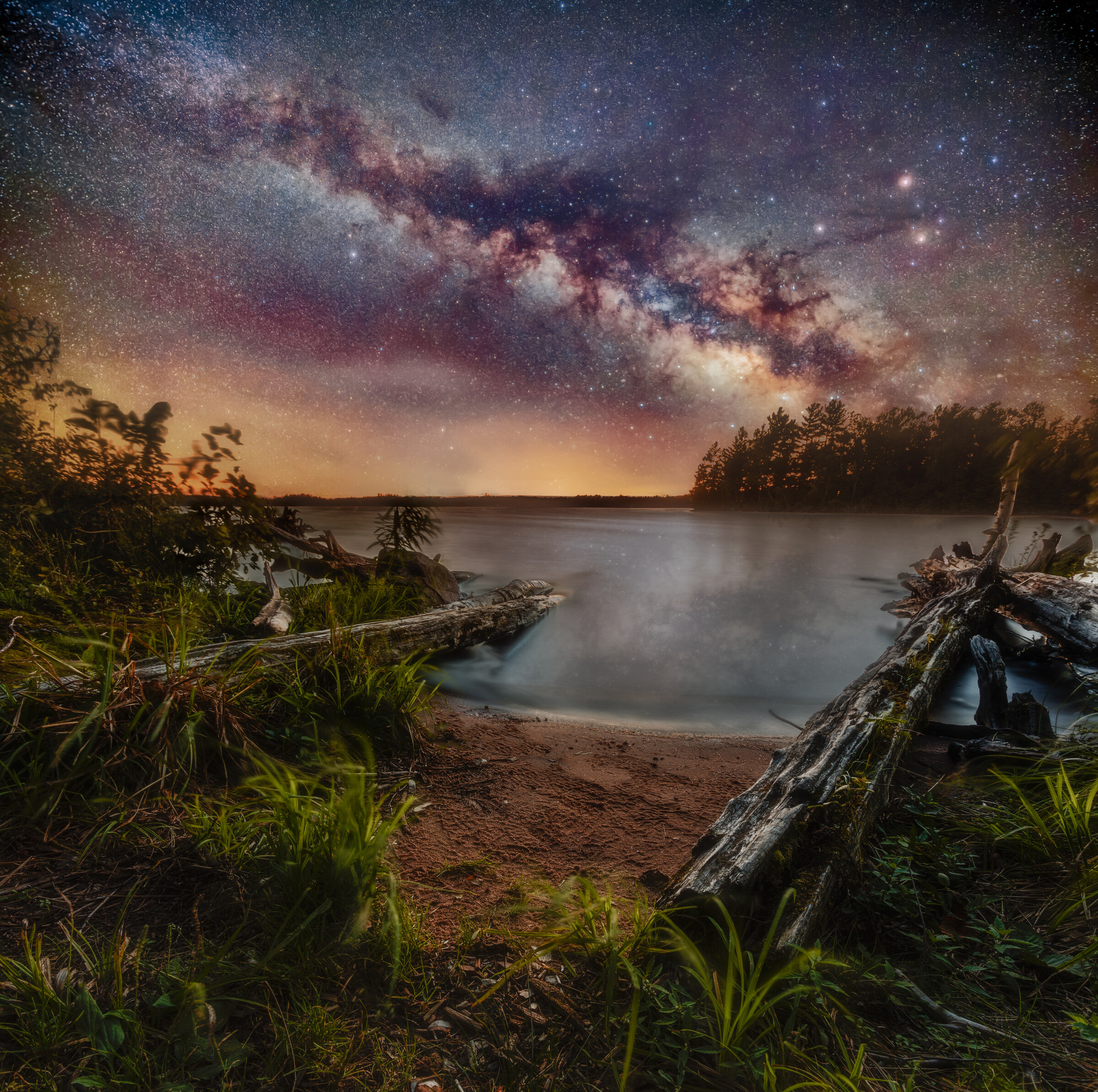 Wide landscape of the Milky Way arching over a quiet inland-sea cove at night, driftwood logs framing a sandy beach and still water