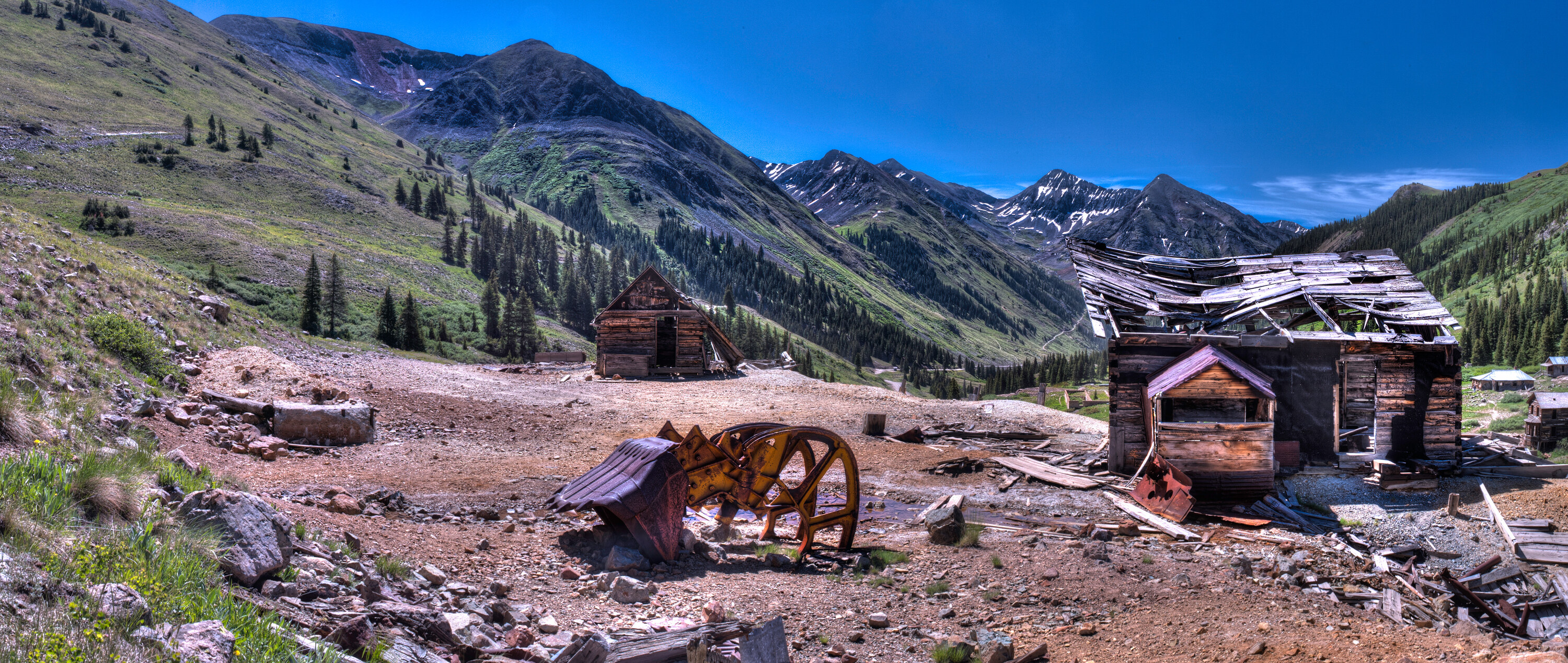 Wide panoramic of the Animas Forks ghost town in the San Juan Mountains, leaning wooden cabins and rusted mining machinery in the foreground, snowy peaks behind