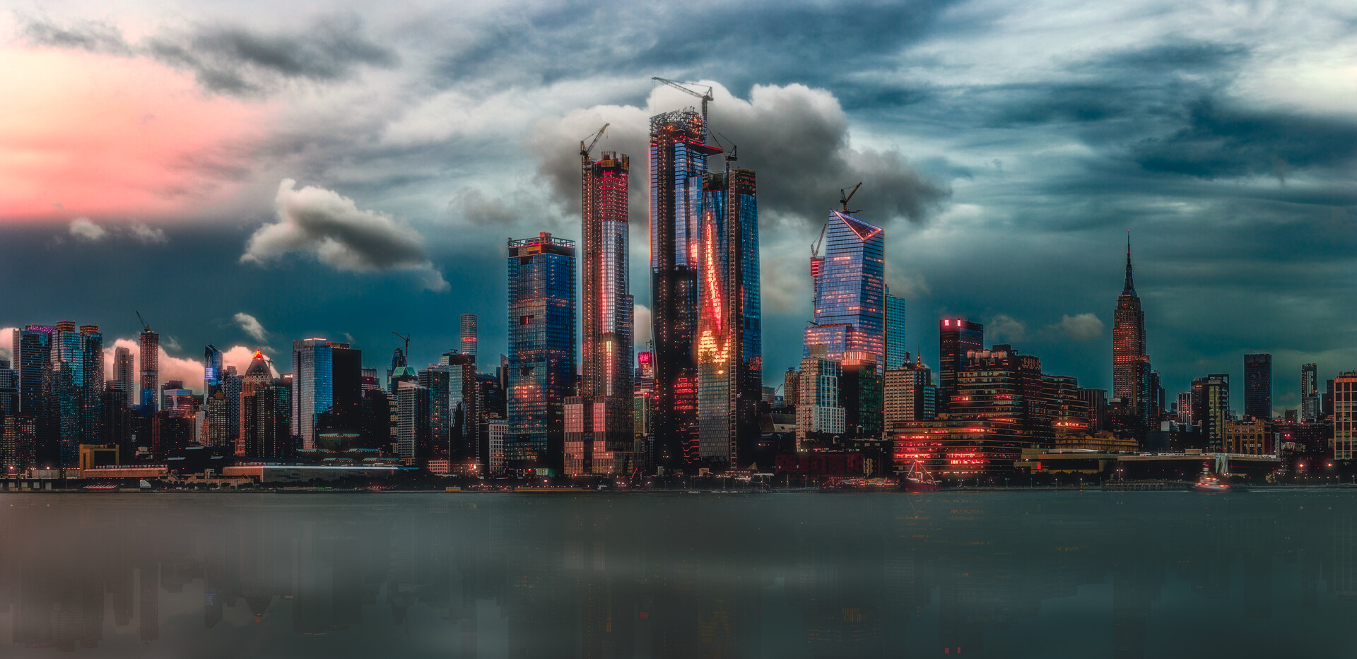Wide panoramic of the Manhattan skyline at twilight with the Hudson Yards towers and construction cranes in the center, the Empire State Building on the right, reflected in the Hudson River