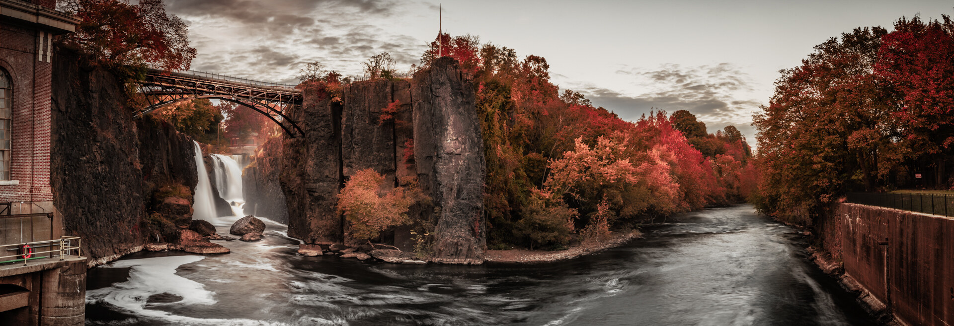 Wide panoramic of Paterson Great Falls in autumn, water dropping over a basalt cliff next to a historic iron bridge, red and orange autumn foliage on both banks