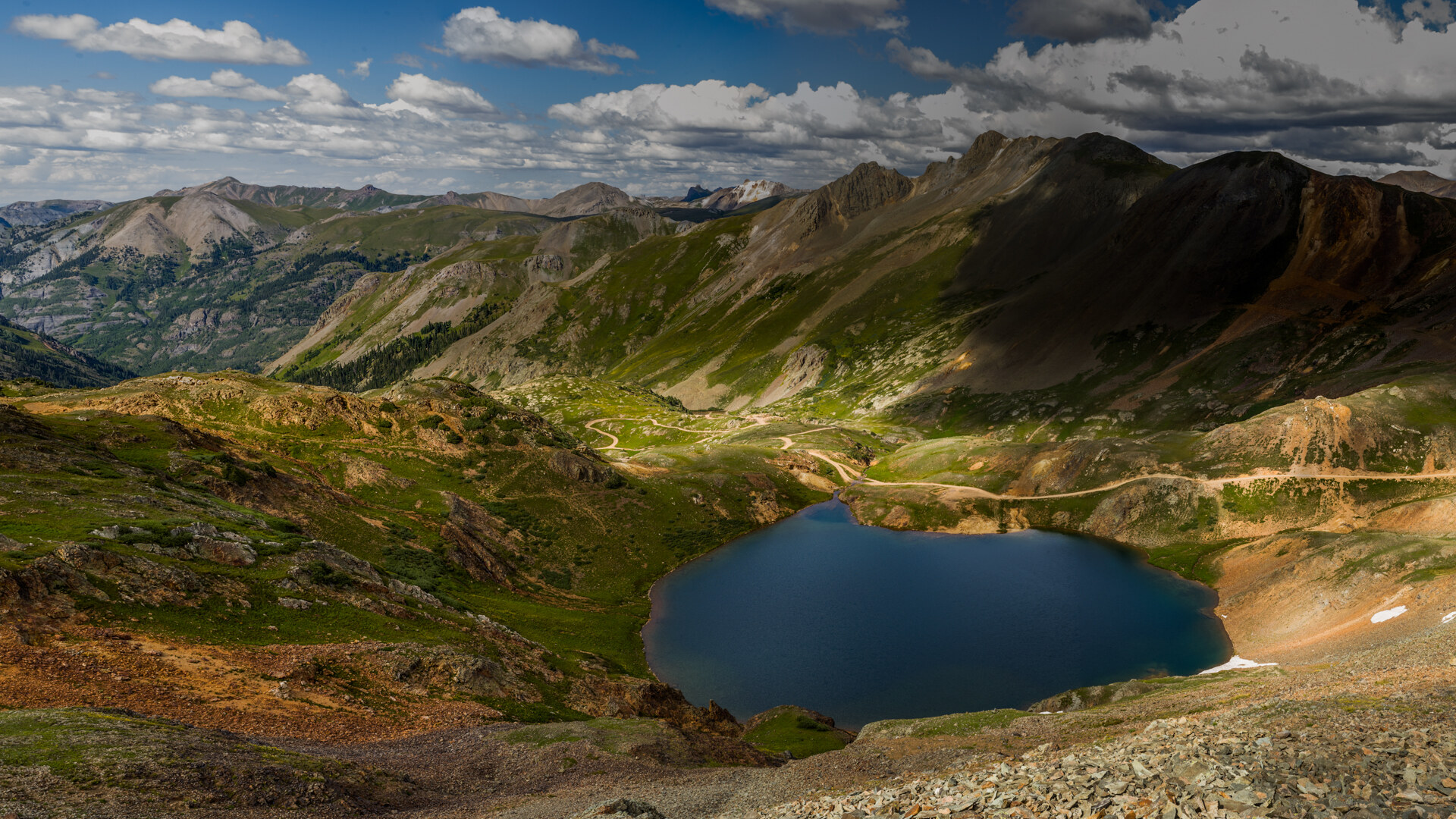 Wide panoramic looking down into a Colorado alpine basin with a turquoise lake at the bottom, a switchback trail winding through the tundra and peaks rising on all sides