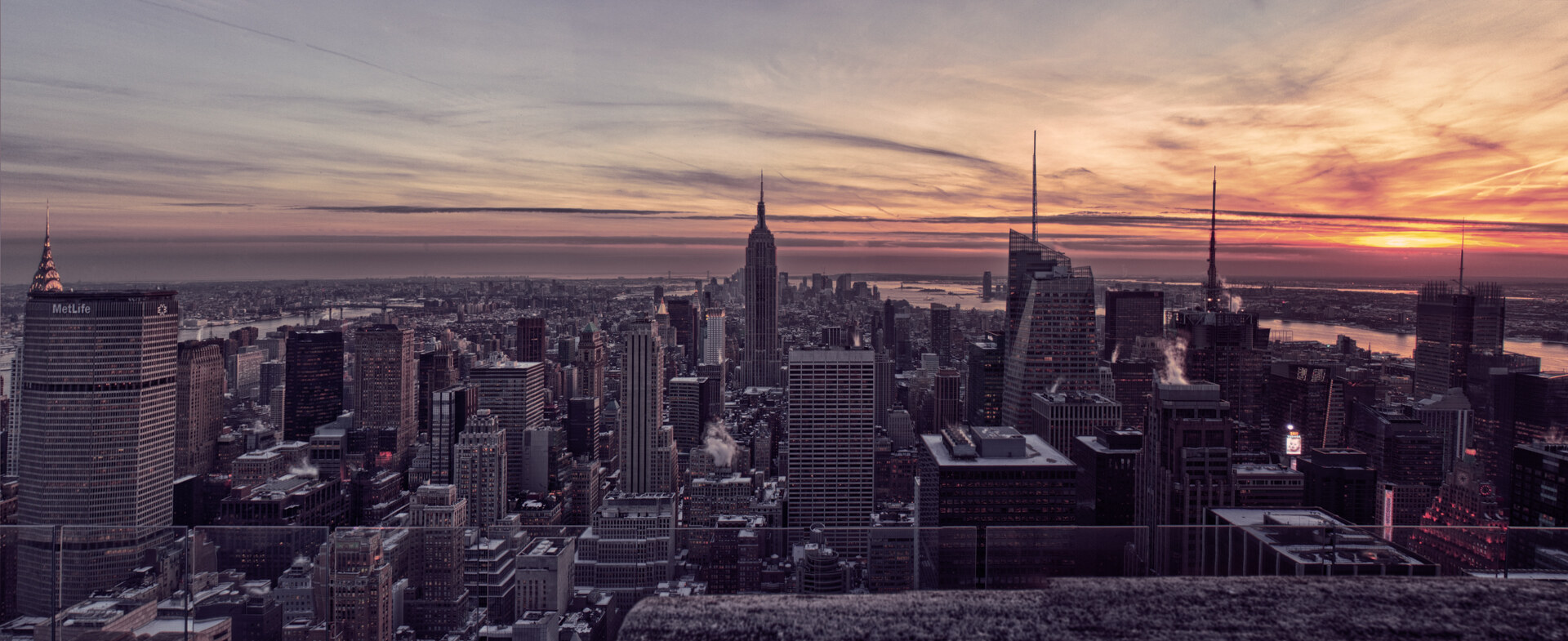 Wide panoramic of the Manhattan skyline from a Midtown rooftop at sunset, the Empire State Building rising in the center, sky in soft pinks and oranges, distant view to the Statue of Liberty