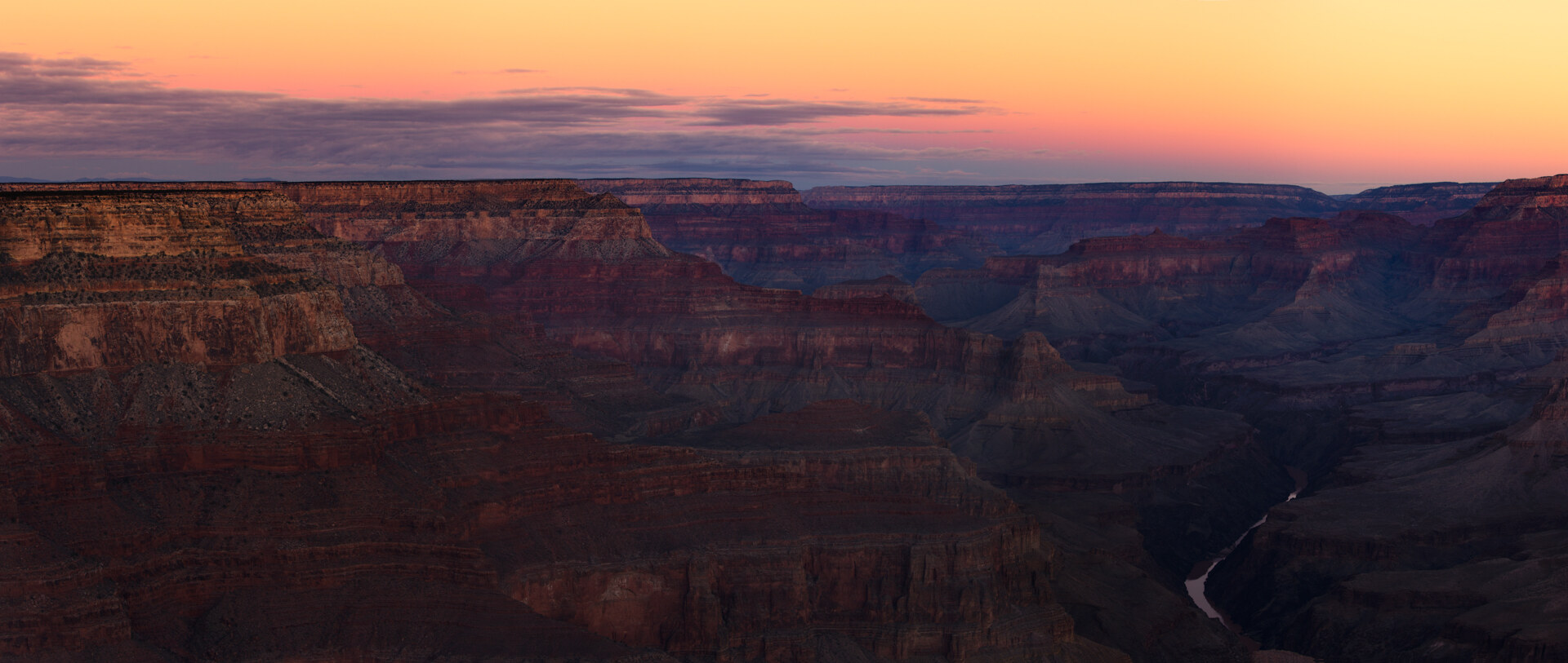 Wide panoramic of the Grand Canyon at sunset, the inner gorge visible on the right and a glimpse of the Colorado River at the bottom, pastel orange-and-purple sky