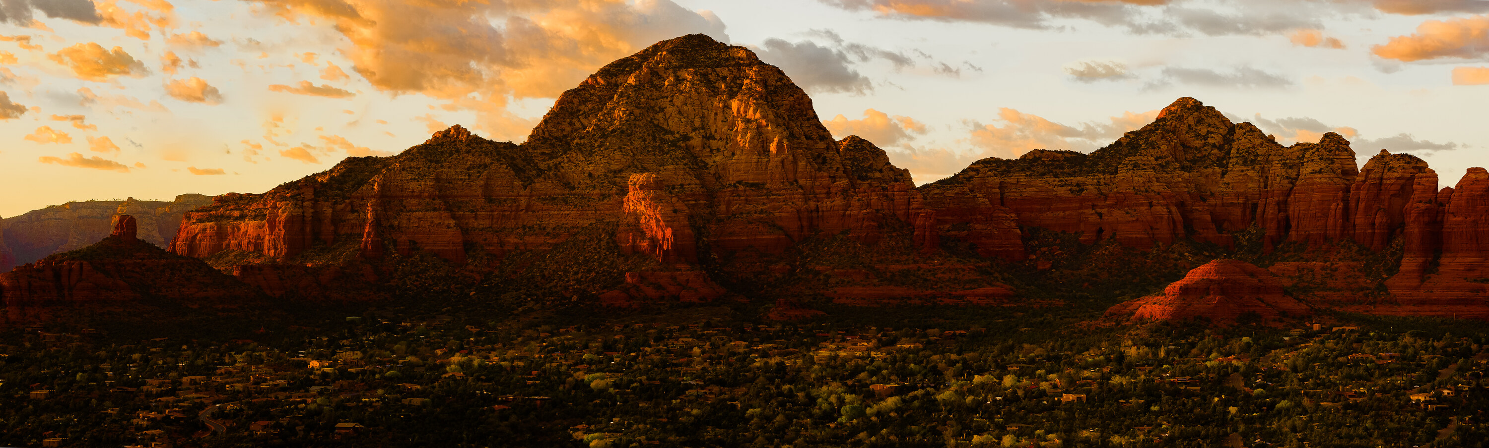 Wide panoramic of the red rocks of Sedona at sunset, Capitol Butte and nearby formations glowing orange-red in side light, the town in shadow below