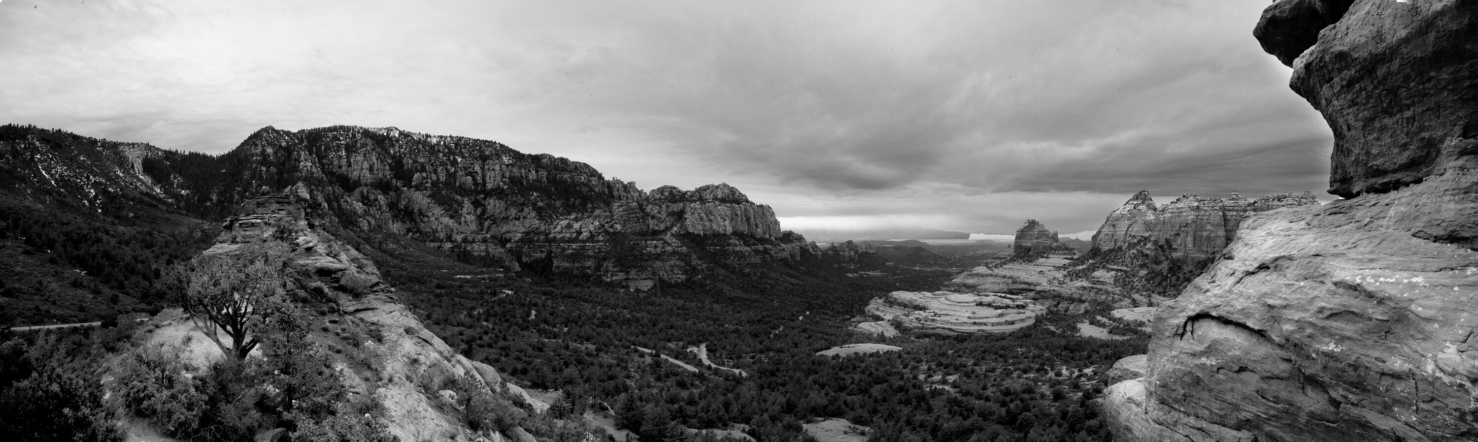 Wide black-and-white panoramic of the Sedona valley with red-rock formations rendered in monochrome, Cathedral Rock in the middle distance and overcast sky above