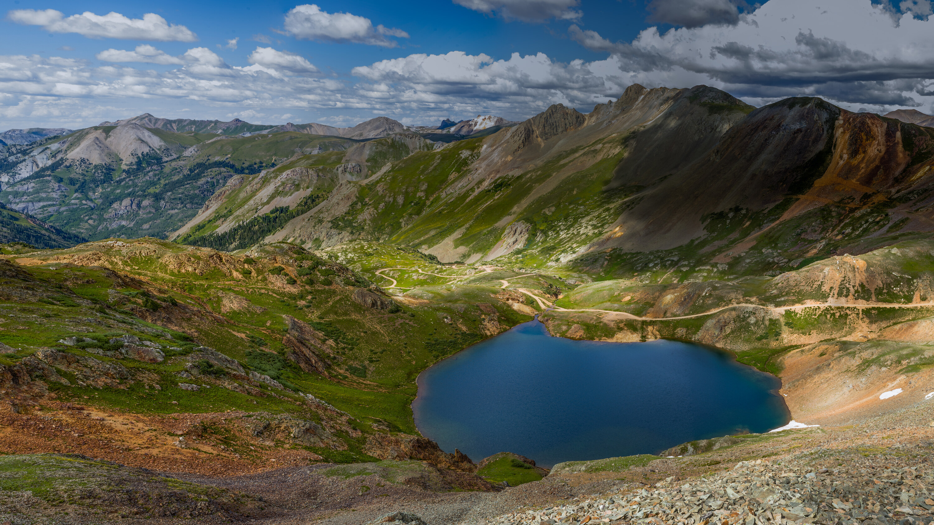 Wide panoramic looking down into a Colorado alpine basin with a deep turquoise lake at the center, surrounding peaks and a trail winding through the valley