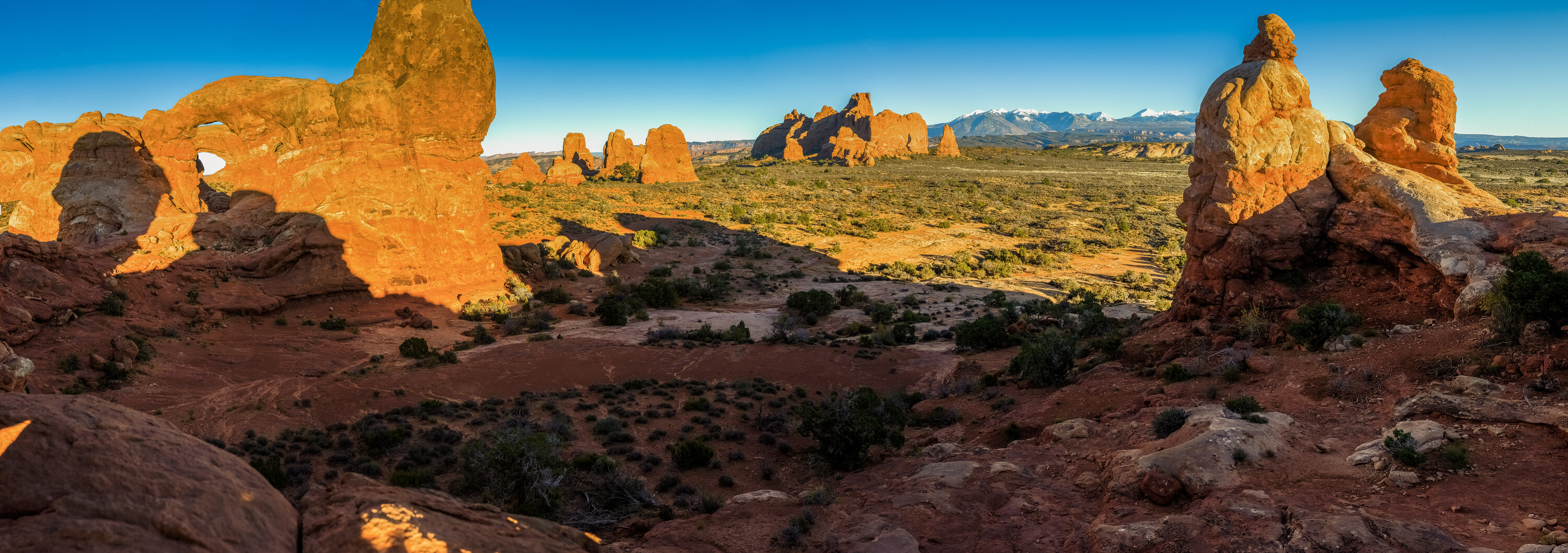 Wide panoramic of the Arches National Park Windows section, Turret Arch visible on the left, sandstone fins across the middle and snow-capped La Sal mountains in the distance