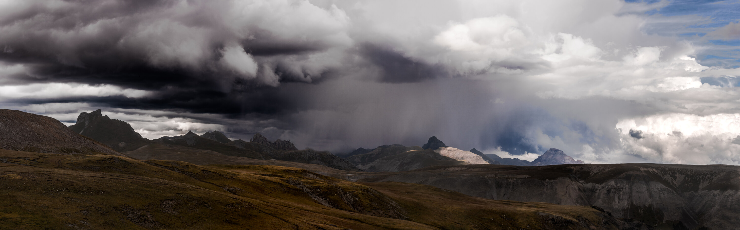 Ultra-wide panoramic of an alpine landscape with a dramatic dark thunderstorm cloud and gray virga rain shaft falling over distant mountain peaks