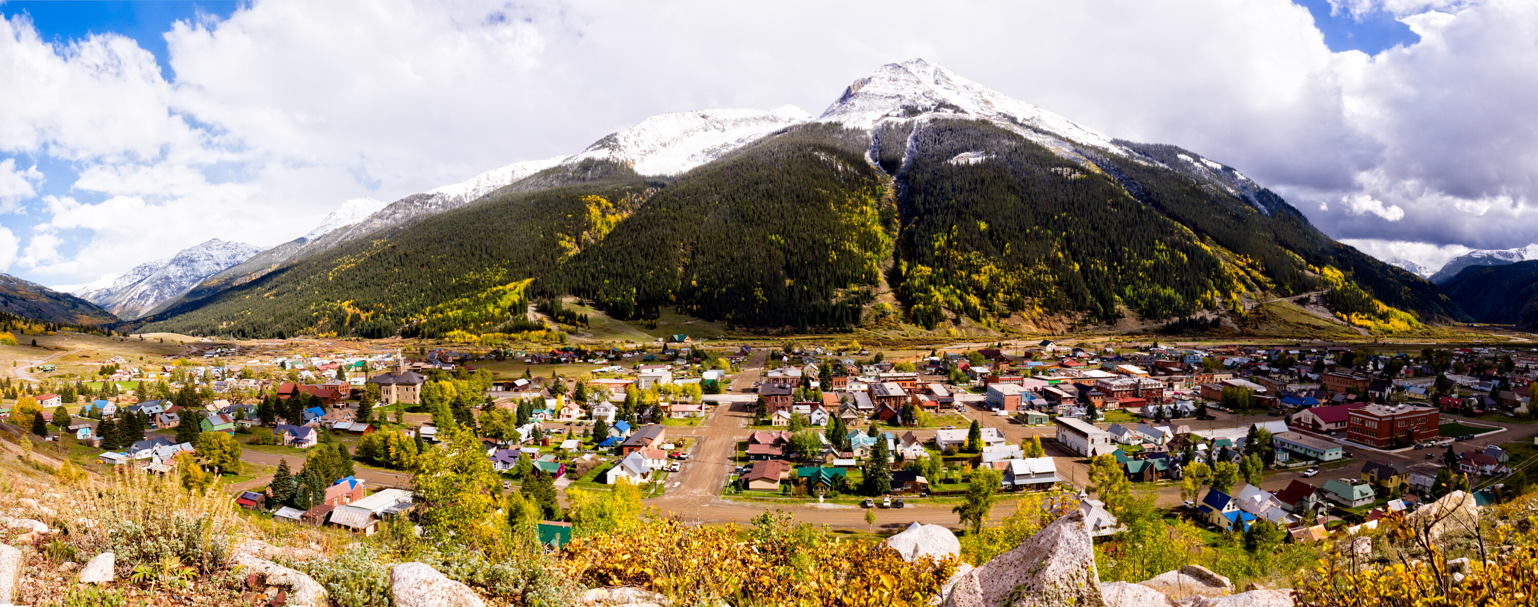 Wide panoramic of the small mountain town of Silverton, Colorado, with snow-dusted peaks rising behind and fall aspen colors on the slopes