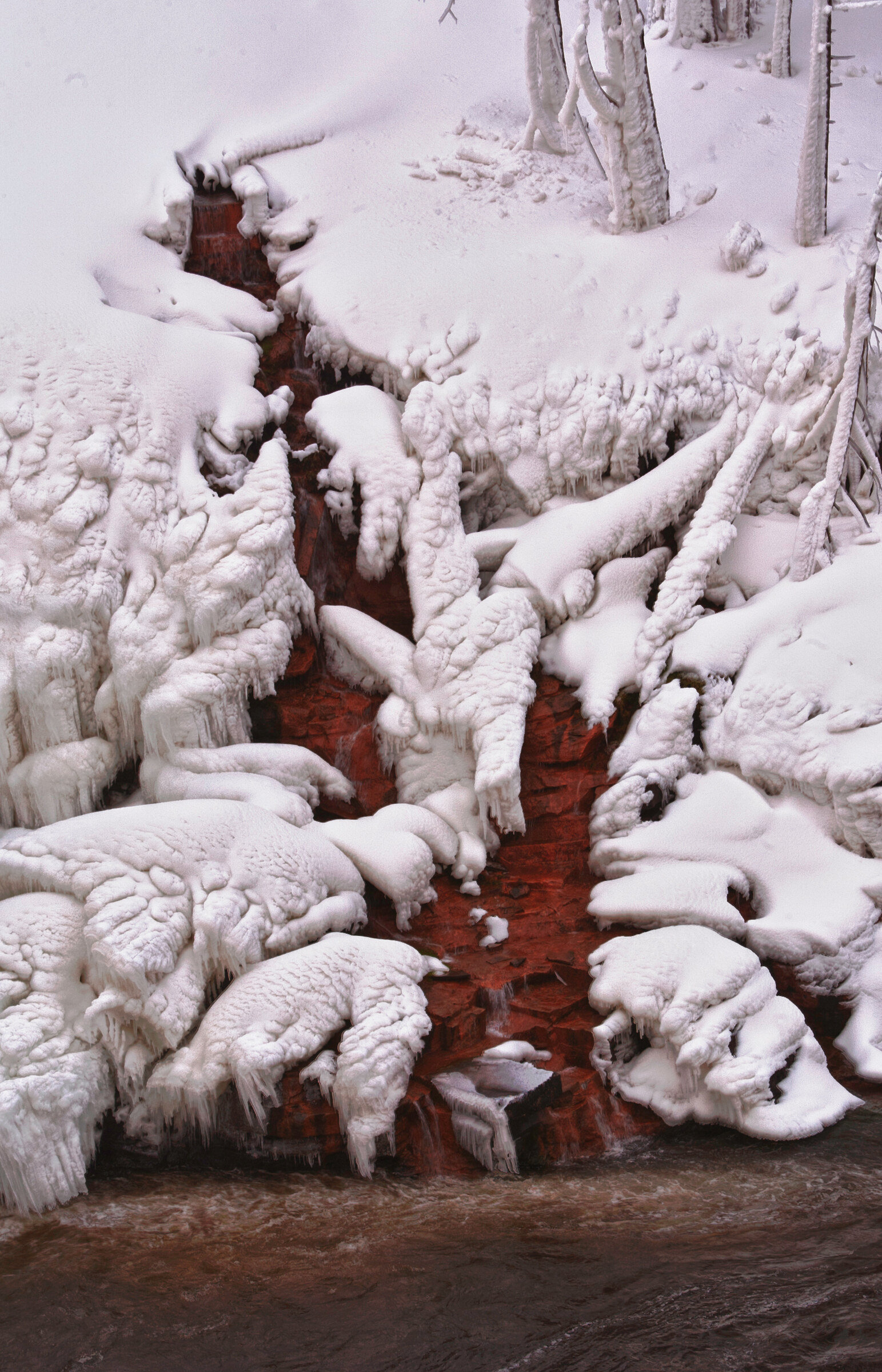 Vertical view of a frozen waterfall over deep red sandstone, thick rope-like ice formations and snow blanketing the entire scene