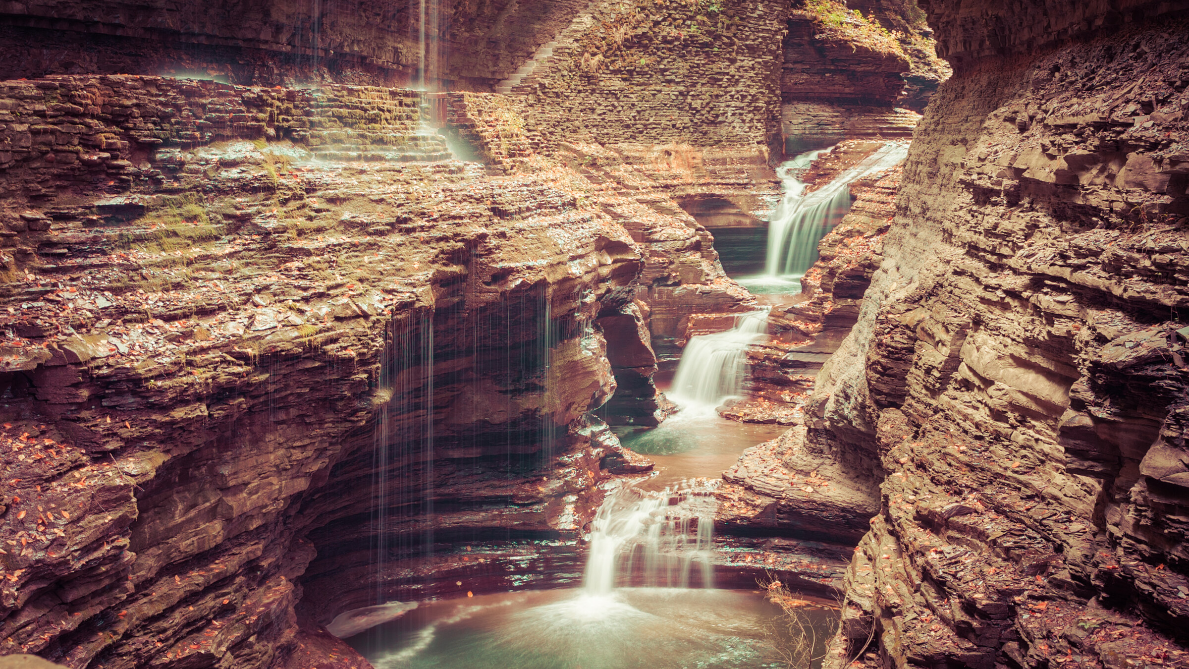 Wide landscape view of Rainbow Falls at Watkins Glen, multiple cascades over deeply layered sandstone and shale, sunlight catching the spray