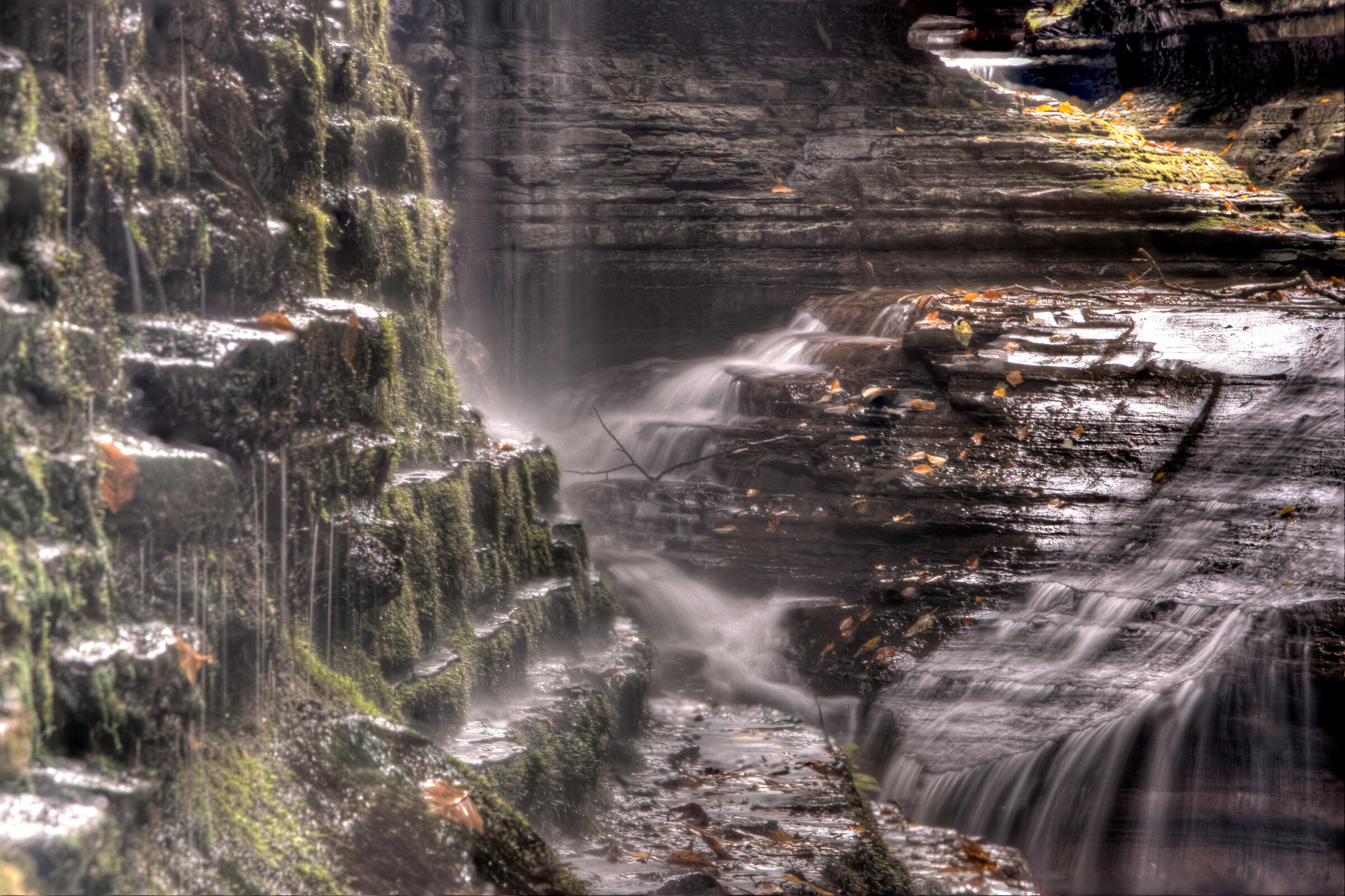 Wide landscape view from behind a curtain of falling water, looking out through the spray at the layered rock walls of Watkins Glen Gorge