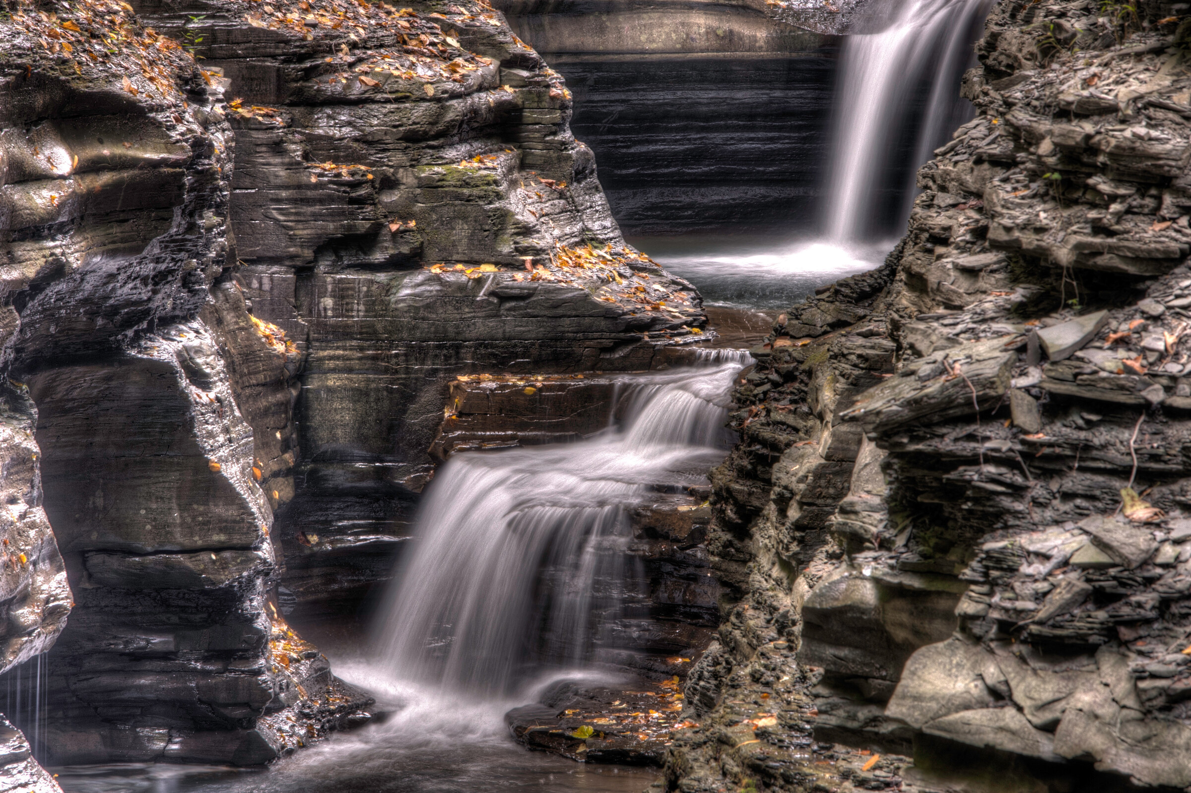 Wide landscape view of a tiered cascade in Watkins Glen, fall leaves resting on smooth wet sandstone ledges, water dropping through multiple pools