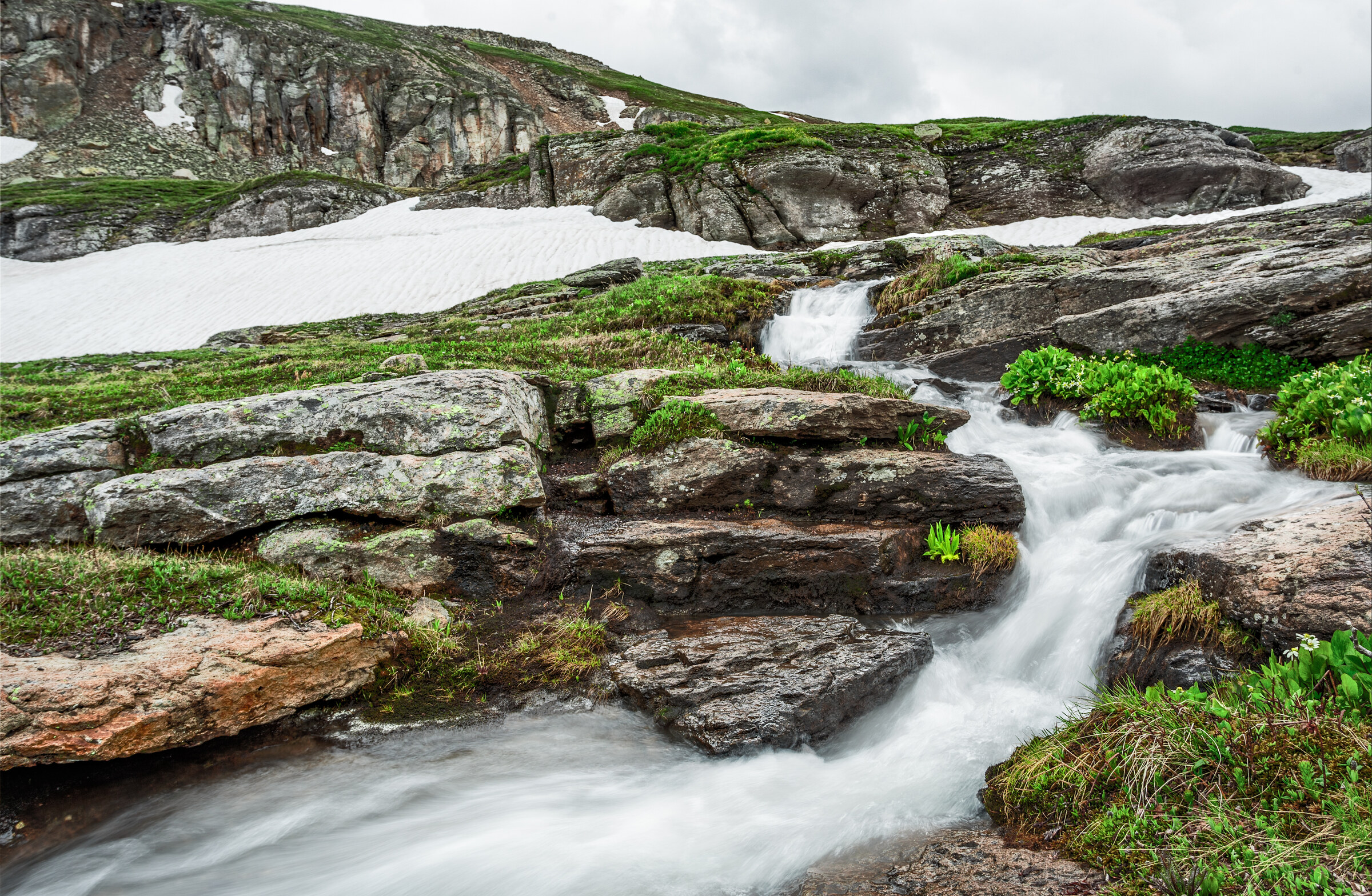 Cascade flowing through tiered granite ledges with snow patches and lush green grass, dramatic cliffs and sky above