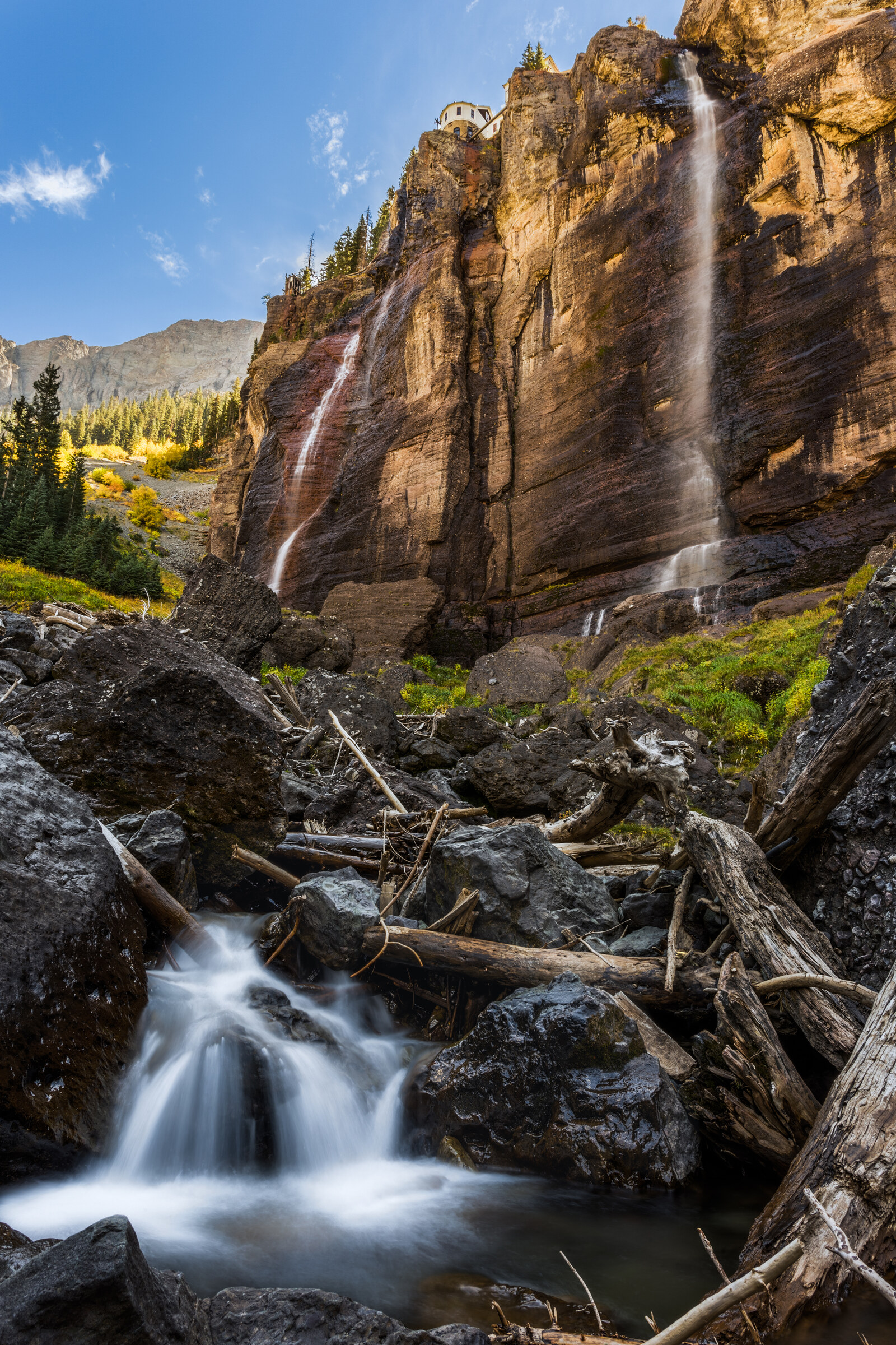 Vertical view of Bridal Veil Falls cascading down a tall red cliff in Telluride, the historic stone power plant at the top, fall colors and a small foreground cascade
