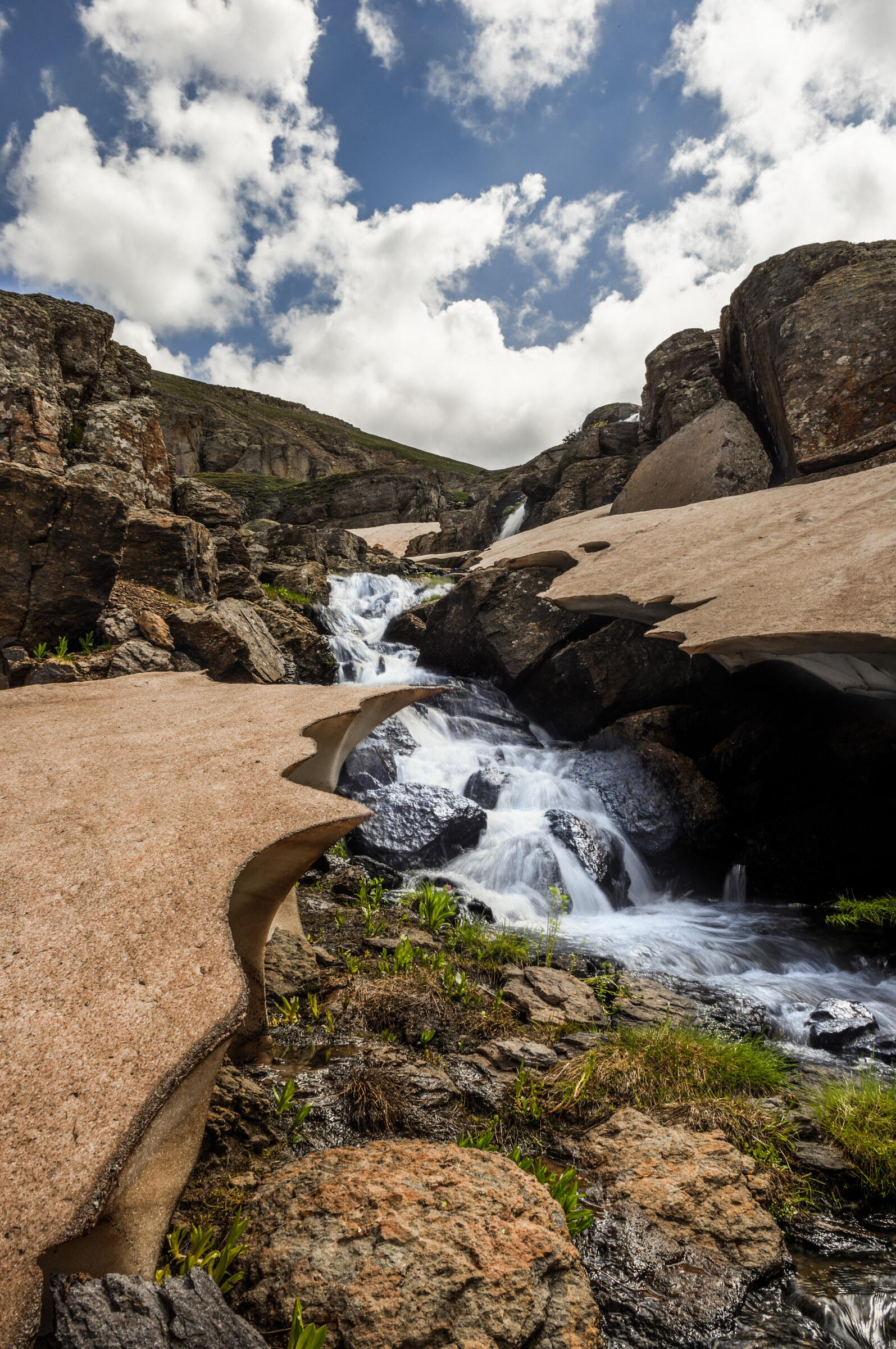 Vertical view of a snowmelt cascade dropping through pink granite ledges, dramatic cumulus clouds above, alpine landscape
